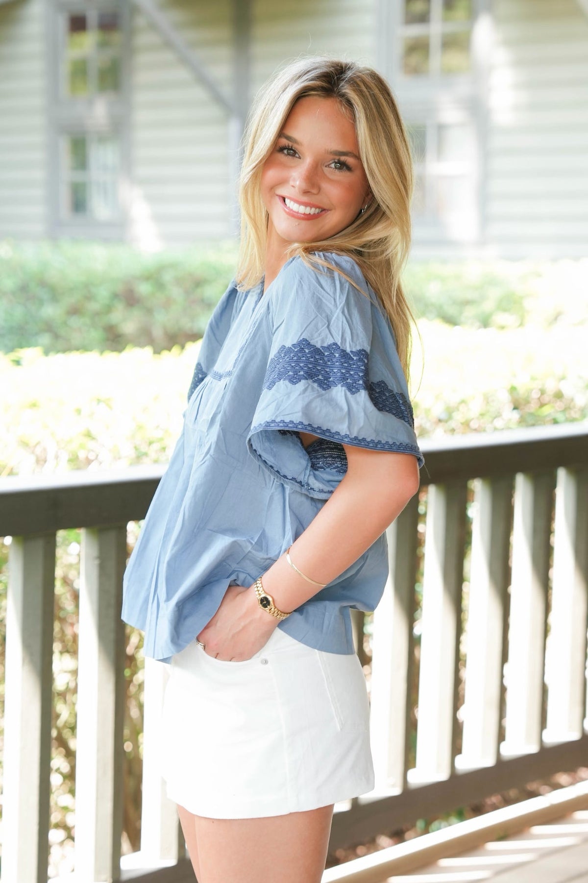 A smiling blonde woman wearing a light blue short-sleeve blouse with dark blue patterned accents and white shorts, standing on a porch with a house and greenery in the background.