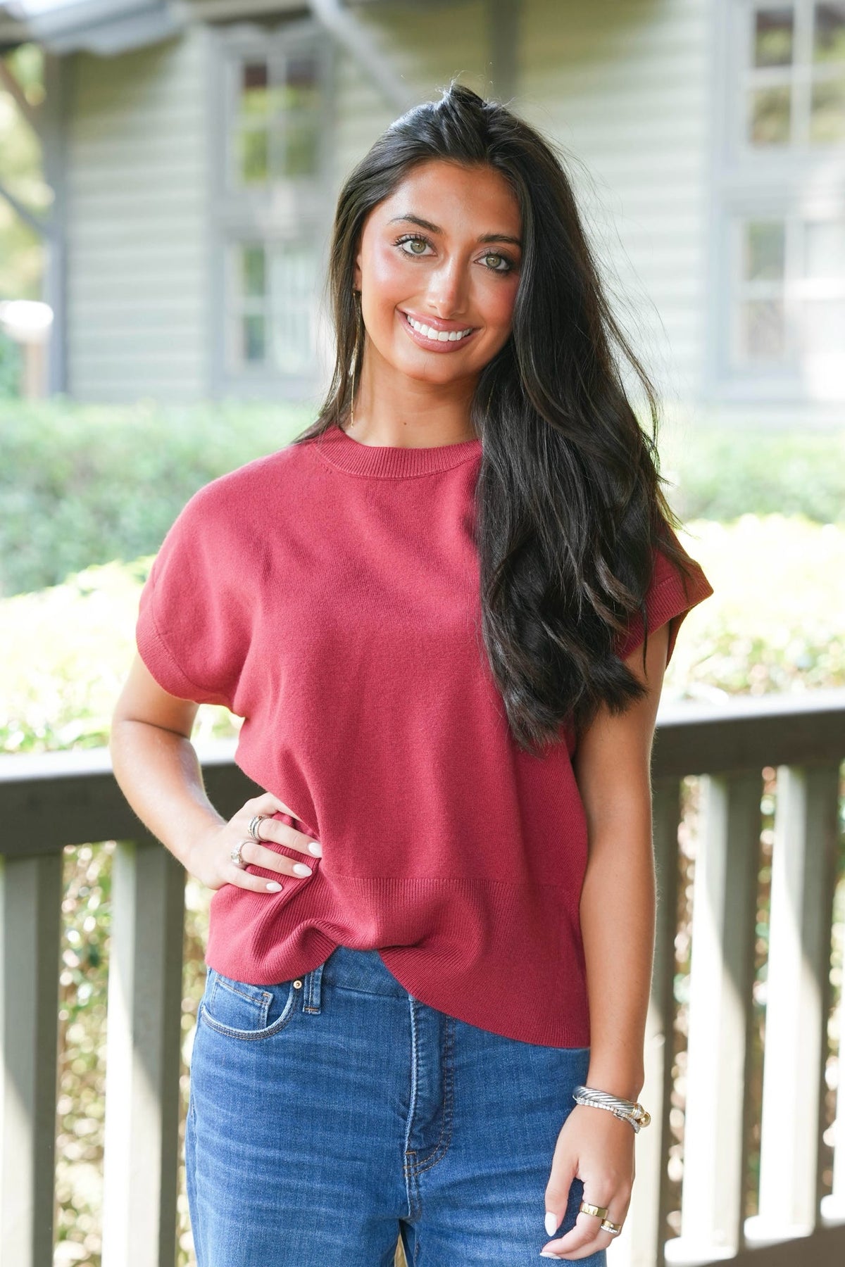 A woman with long dark hair stands on a porch wearing a loose-fitting, short-sleeve crewneck t-shirt in a dusty rose red color, paired with blue jeans.