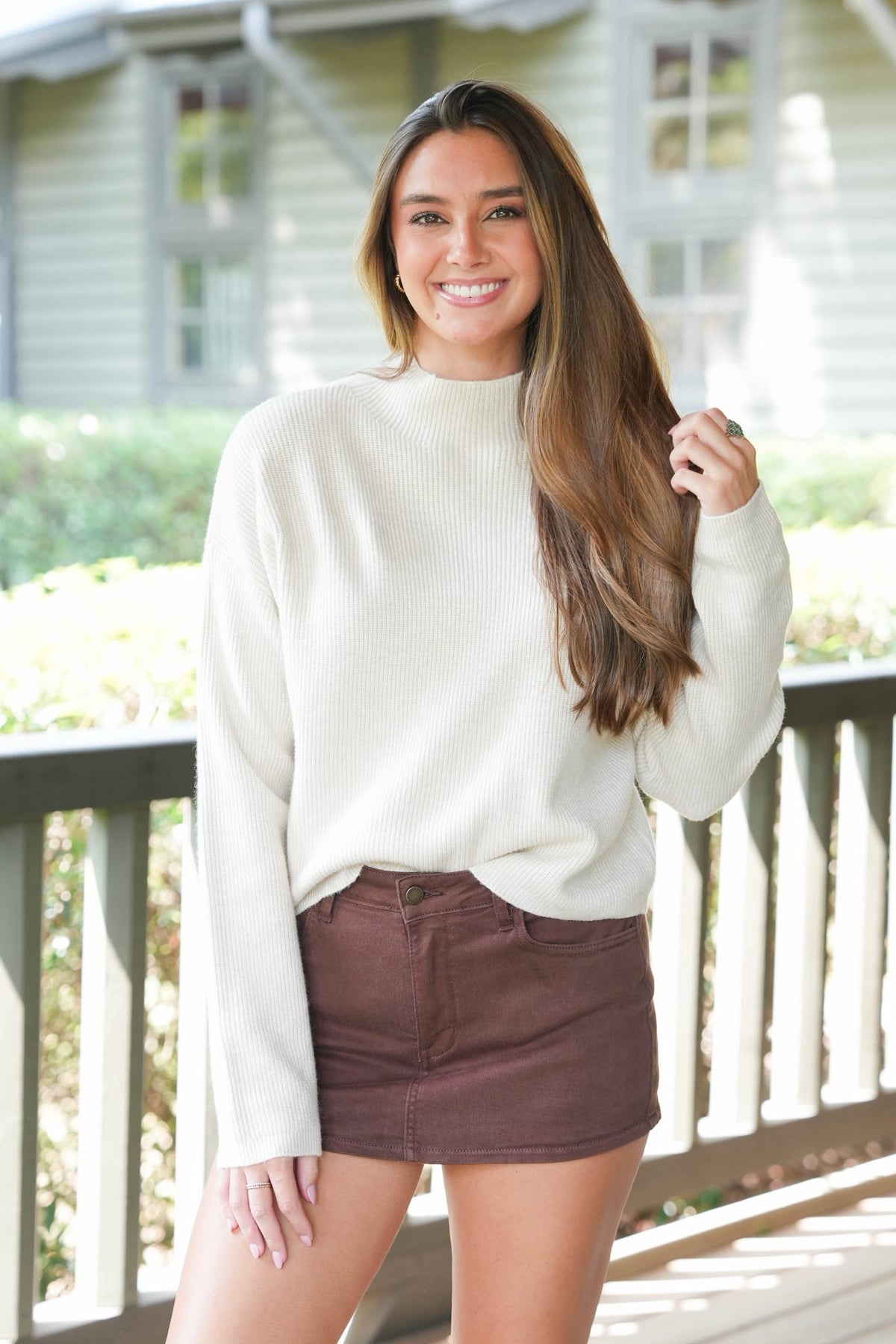 A woman smiling on an outdoor porch, wearing a cream ribbed mock neck sweater and a brown mini skirt.