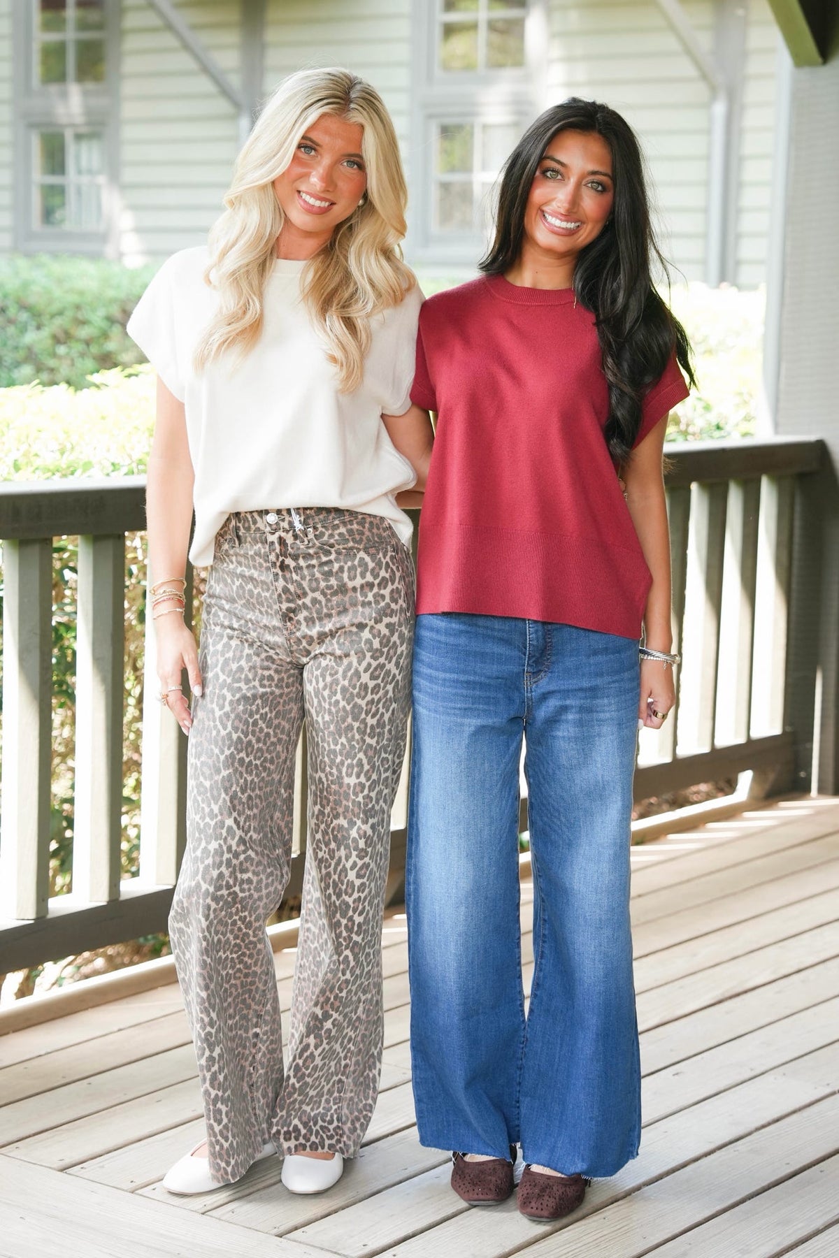 Two women standing together on a wooden deck wearing casual summer outfits. The woman on the left has blonde hair and wears a white short-sleeve top with leopard-print wide-leg pants.