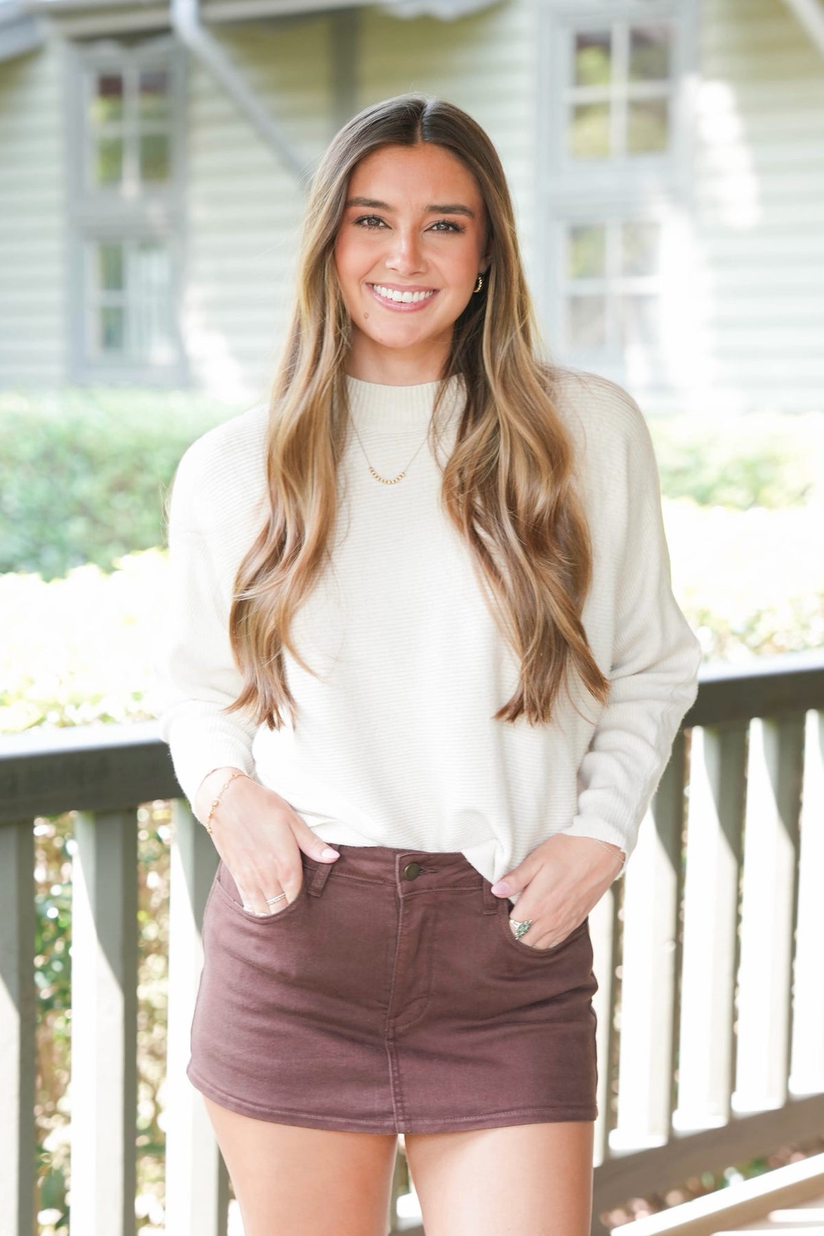 A smiling woman with long light brown hair stands on a porch wearing a cream mock-neck knit sweater and a brown mini skirt.