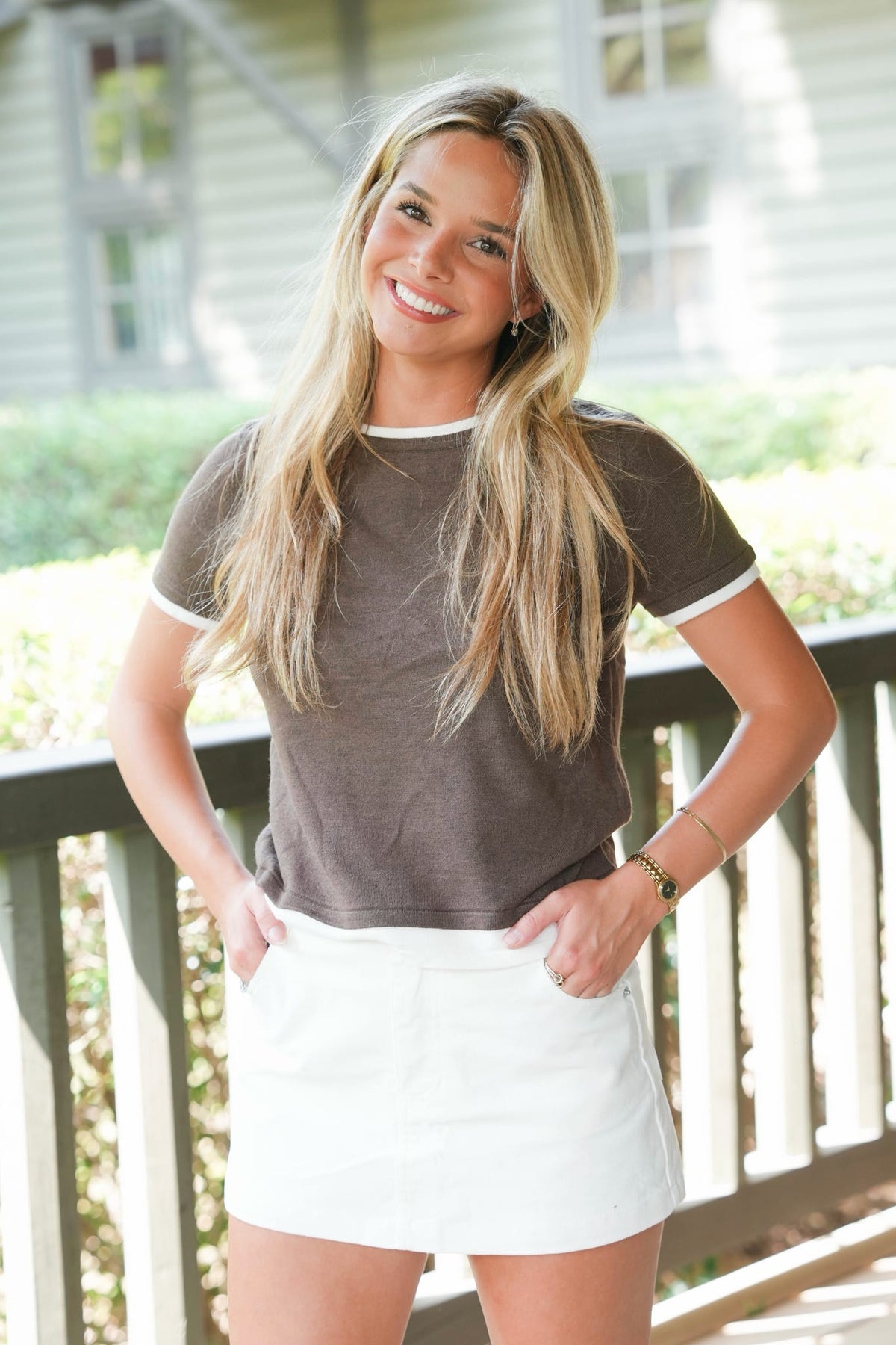 A smiling blonde woman wearing a brown ringer t-shirt with white trim and a white mini skirt, standing on a wooden porch with a house in the background.