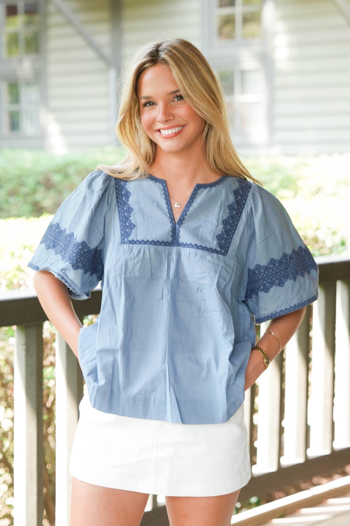A smiling blonde woman wearing a light blue short-sleeve blouse with dark blue embroidered trim and a white skirt, standing on a porch with a house in the background.