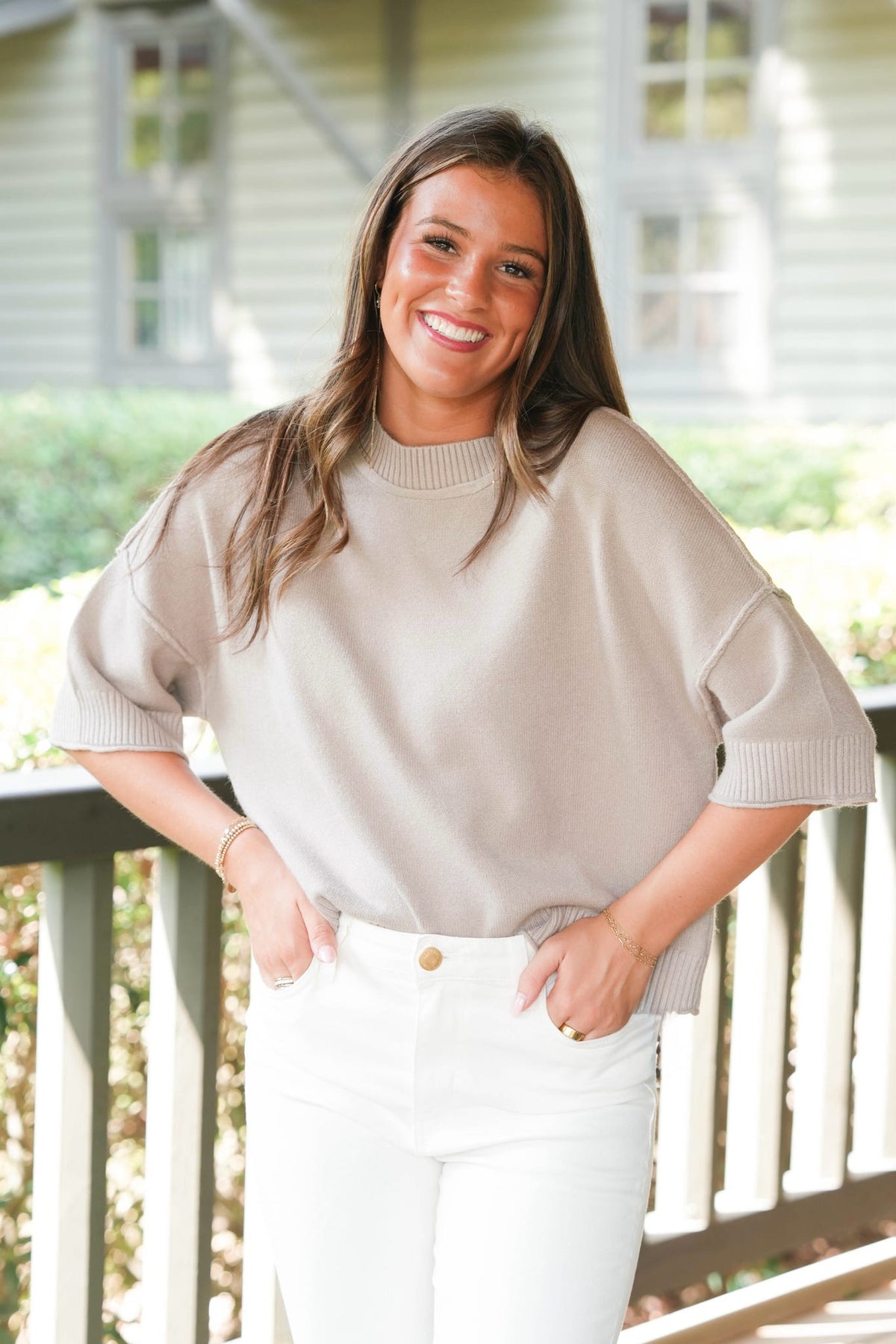 A smiling woman wearing a beige short-sleeve knit sweater and white pants, standing on a porch with a house in the background.