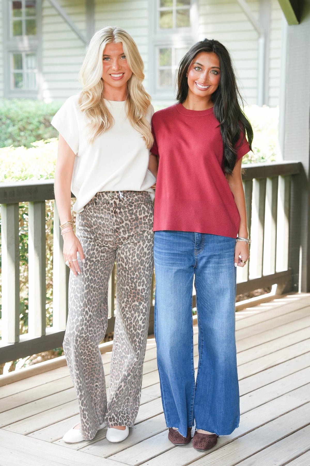 Two women standing together on a wooden deck outdoors. The woman on the left wears a white short-sleeve top and leopard-print wide-leg pants, while the woman on the right wears a dusty red short-sleeve top and blue wide-leg jeans.