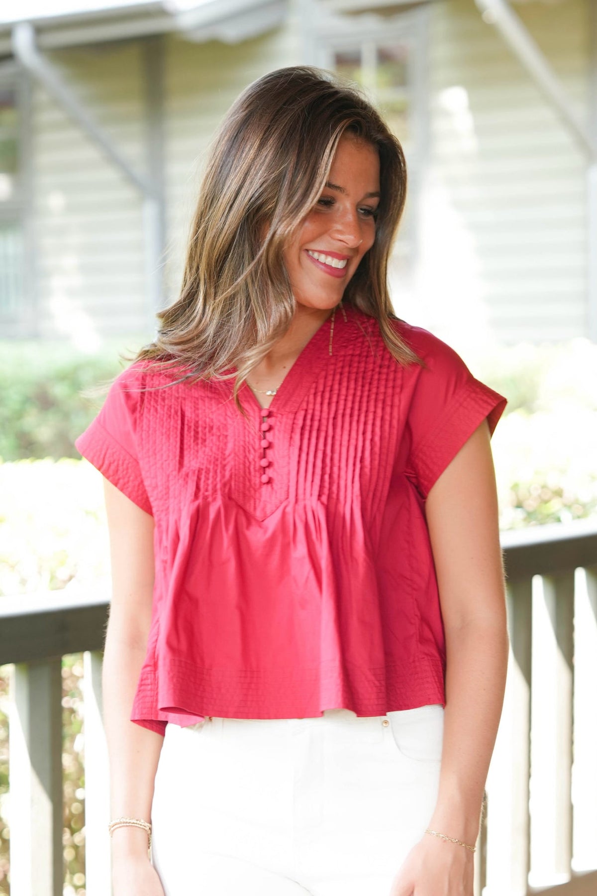 A woman smiling on a porch wearing a bright pinkish-red short-sleeve top with vertical pleats and white pants.