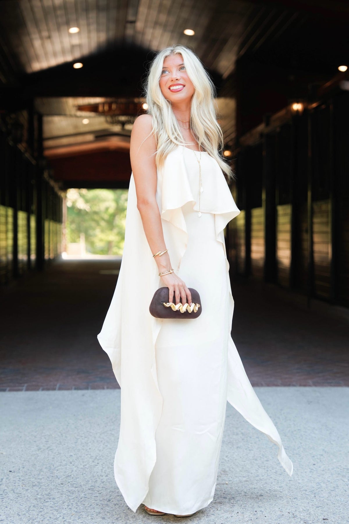 A woman with long blonde hair stands in a covered walkway wearing a sleeveless, off-white ruffled maxi dress and holding a small dark brown clutch with gold hardware.