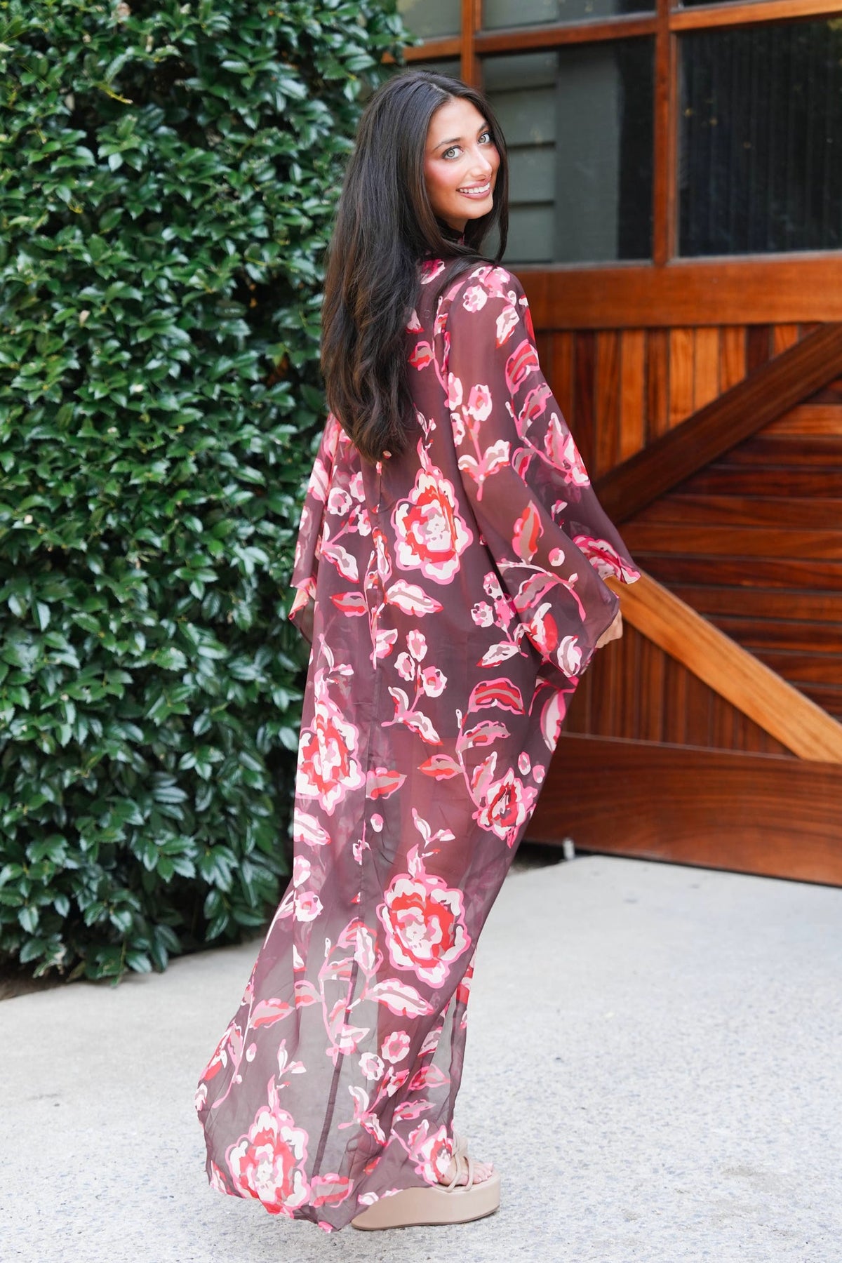 A woman wearing a long, sheer mauve-brown kaftan dress with a red and pink floral print, standing outdoors in front of a wooden barn door and green bushes.