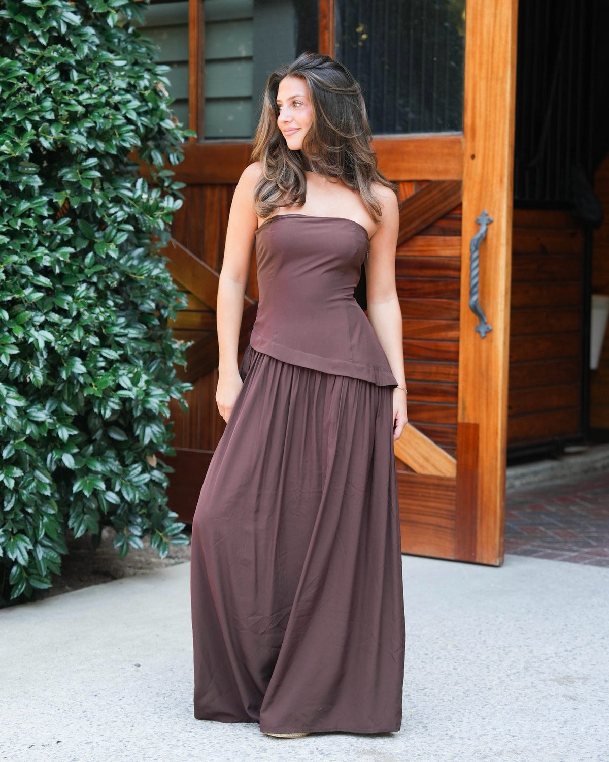 A woman wearing a two-piece brown dress standing in front of a wooden door and green bushes.