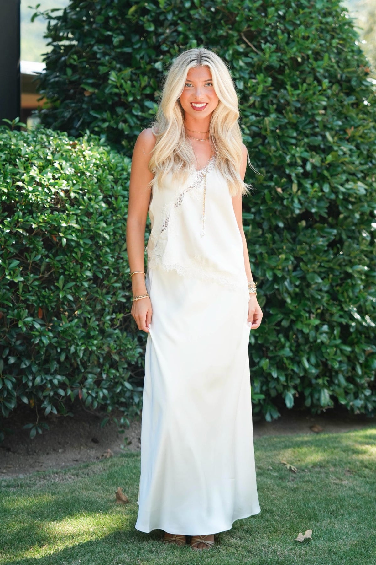 A woman with long blonde hair stands outdoors in a garden wearing a sleeveless, floor-length ivory slip dress with delicate beaded trim along the neckline. The background consists of green hedges and grass.