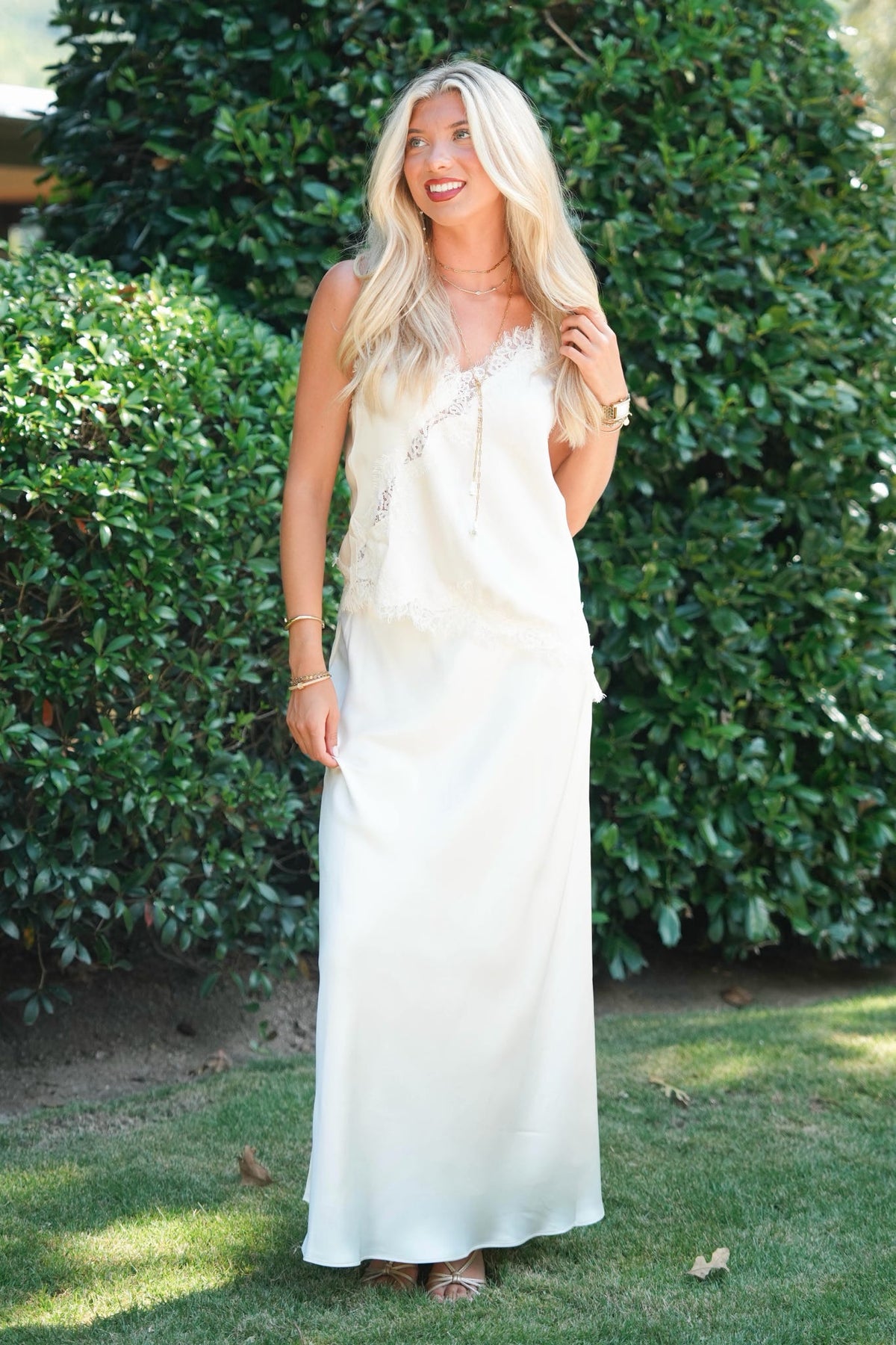 A woman wearing a long cream satin slip dress with lace-up detailing stands outdoors in a grassy garden area with green hedges behind her.