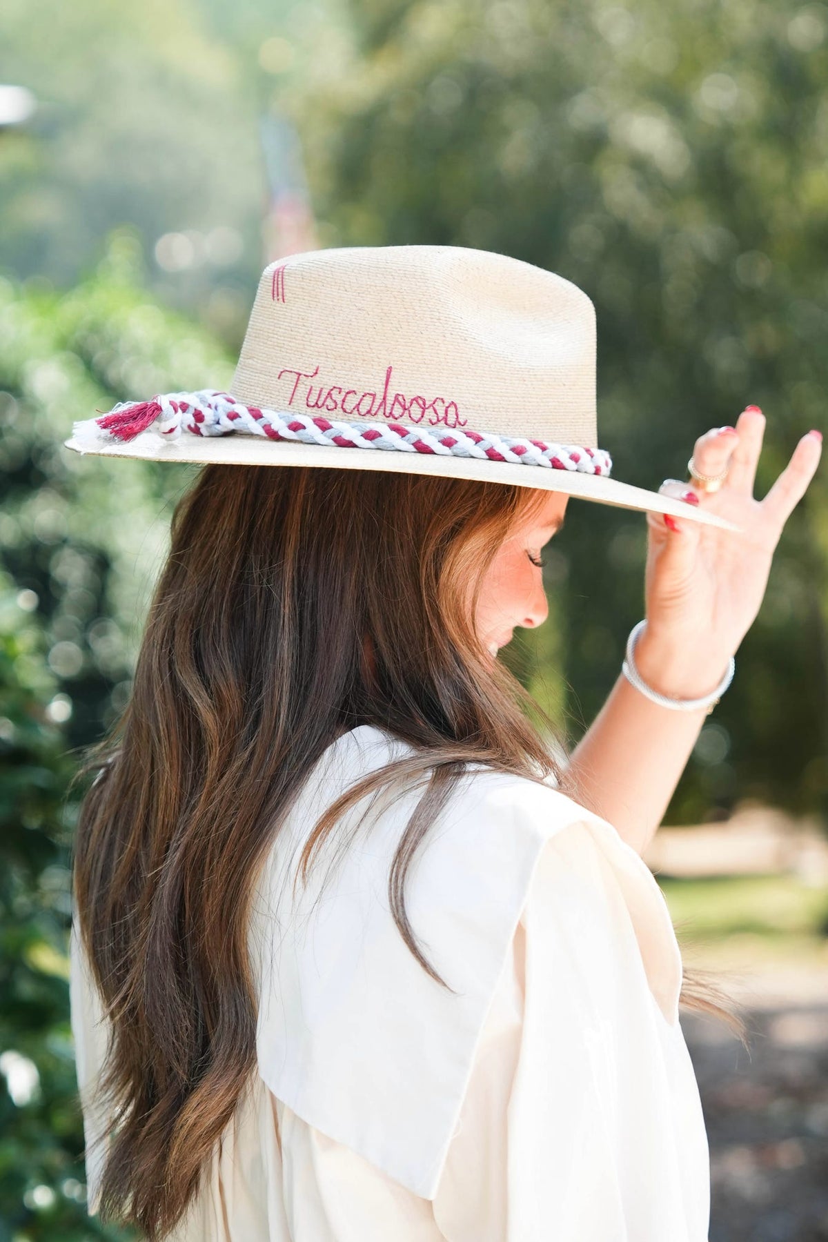 A woman wearing a beige wide-brim fedora hat with 'Tuscaloosa' embroidered on the crown and a red-and-white patterned band, photographed outdoors against a blurred green background.