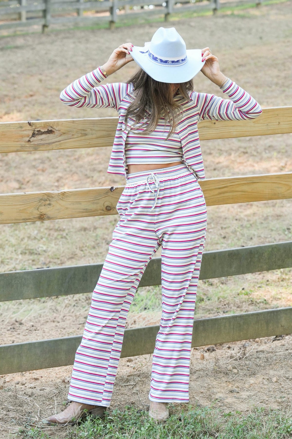 A woman wearing a matching two-piece striped lounge set with a white cowboy hat, posing outdoors in front of a wooden fence.