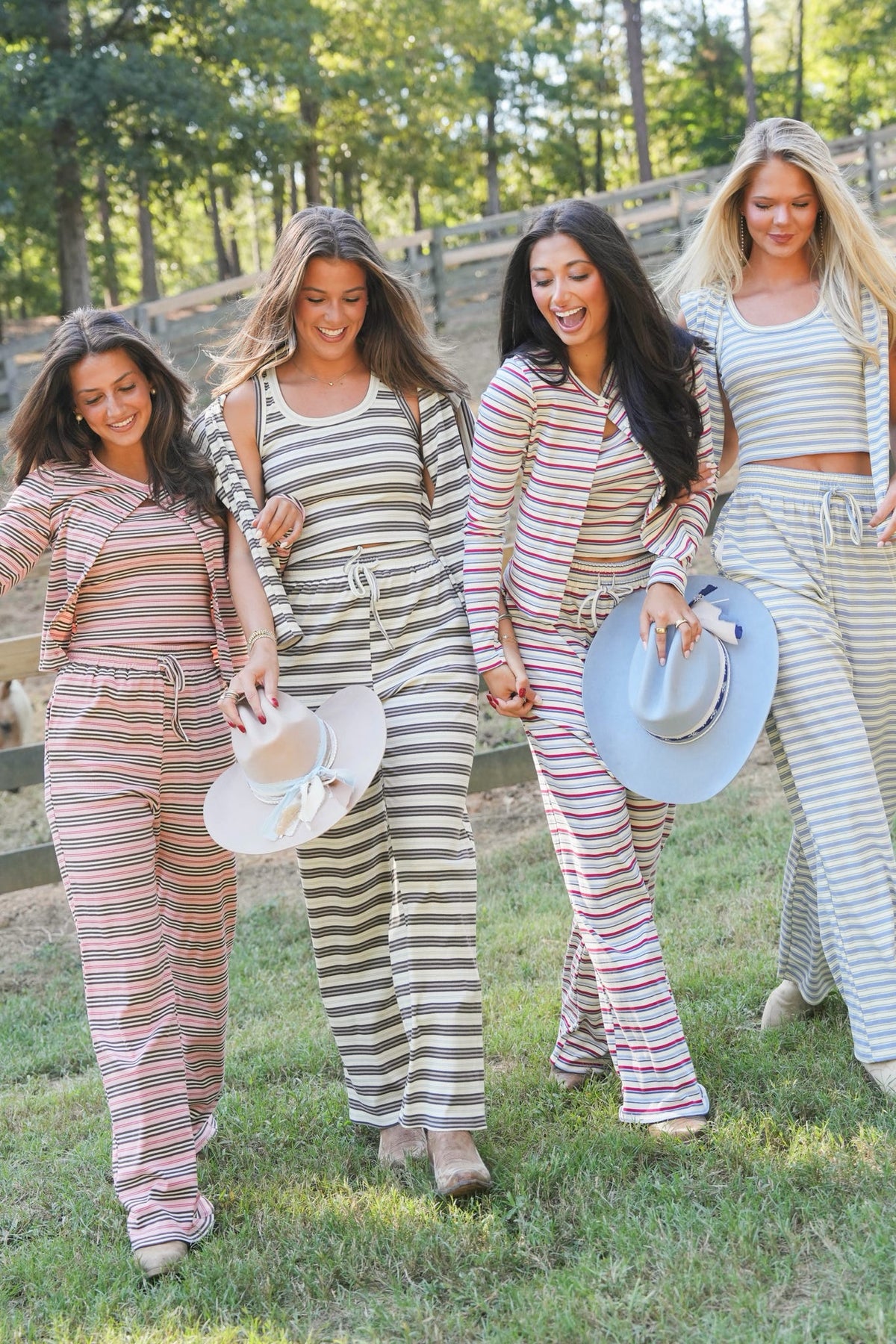Four women walking outdoors on a grassy area with trees and a wooden fence in the background, wearing matching striped lounge sets in different colorways. One woman holds a white hat with a bow, another holds a light blue hat.