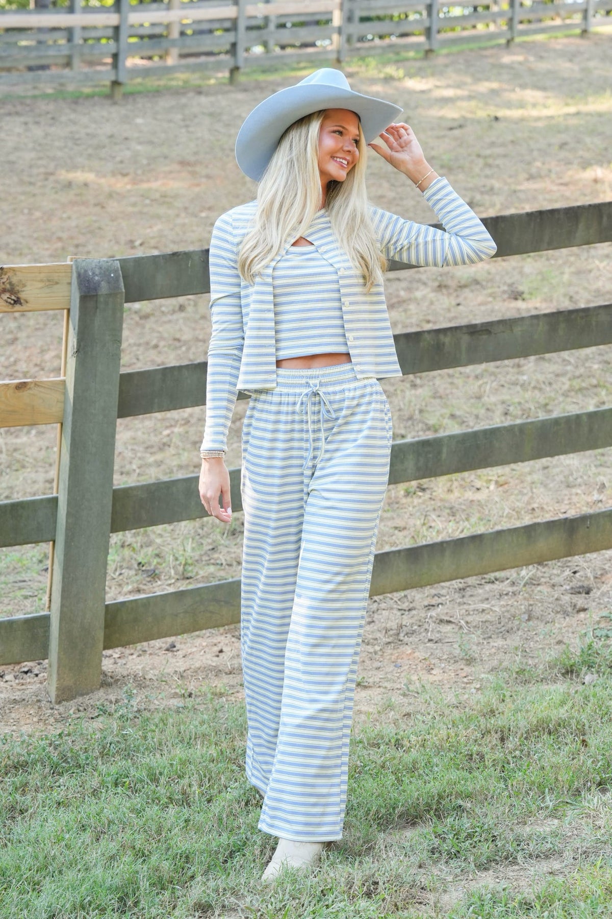 A woman stands outdoors in front of a wooden fence wearing a light blue and white striped long-sleeve crop top and matching wide-leg pants, along with a wide-brimmed light blue hat.