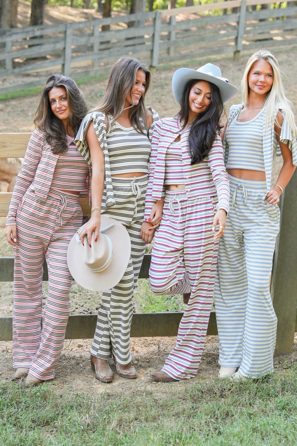 Four women posing together outdoors in matching striped two-piece lounge sets with wide-leg pants and crop tops, standing in front of a wooden fence.