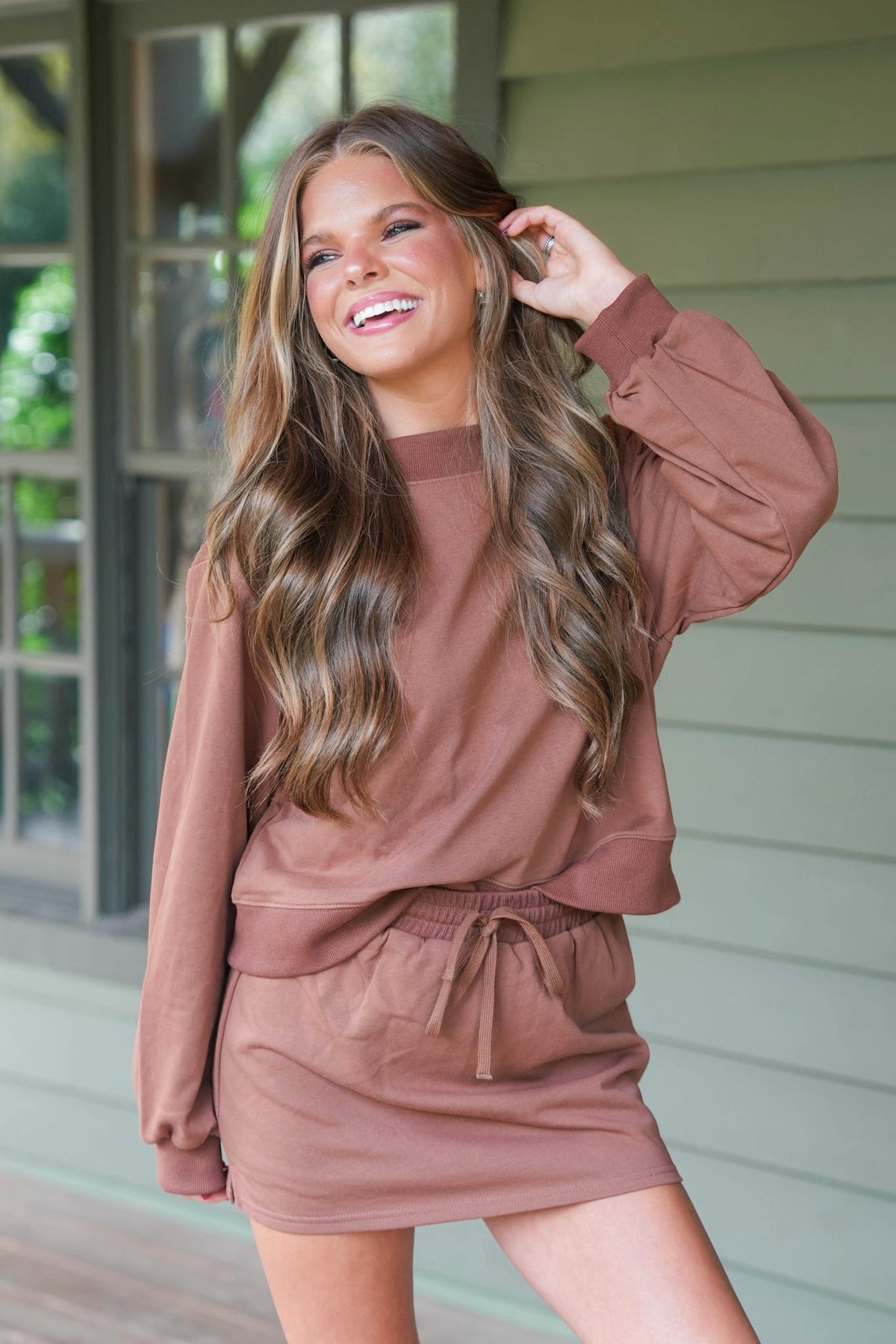 woman standing on a wooden porch wearing a brown matching sweatshirt and skort set
