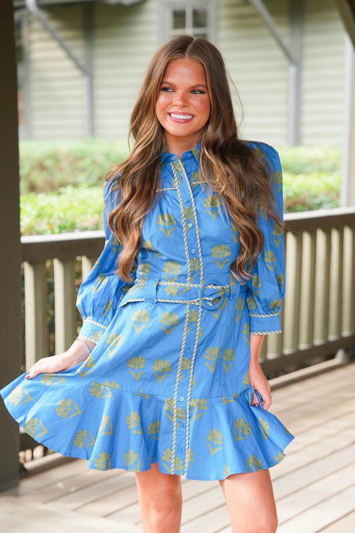 A woman modeling a light blue long-sleeve mini dress with a yellow floral pattern and gold trim, standing on a wooden porch.