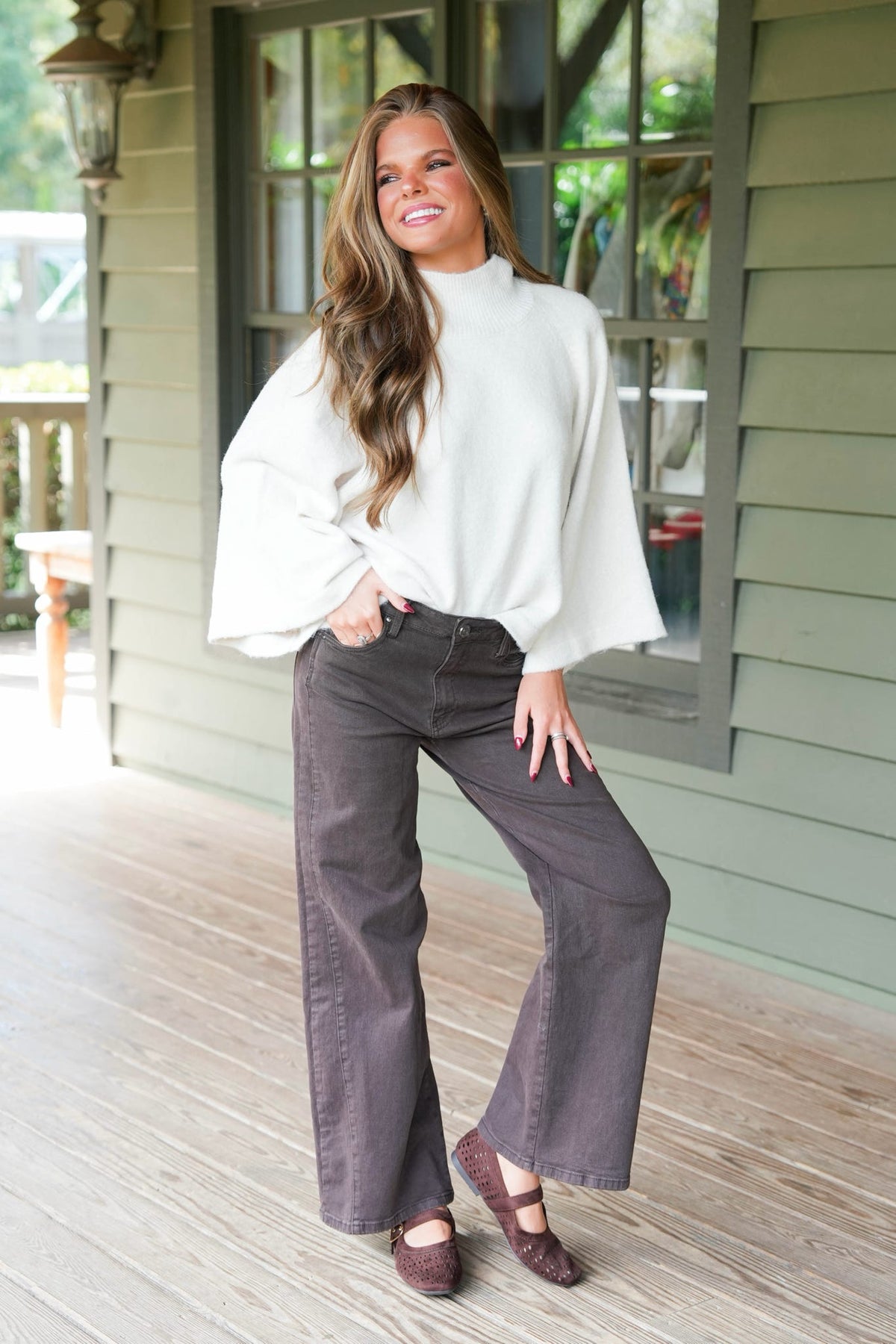 A woman posing on a wooden porch wearing a white mock-neck long-sleeve top, dark gray wide-leg pants, and brown flat shoes.
