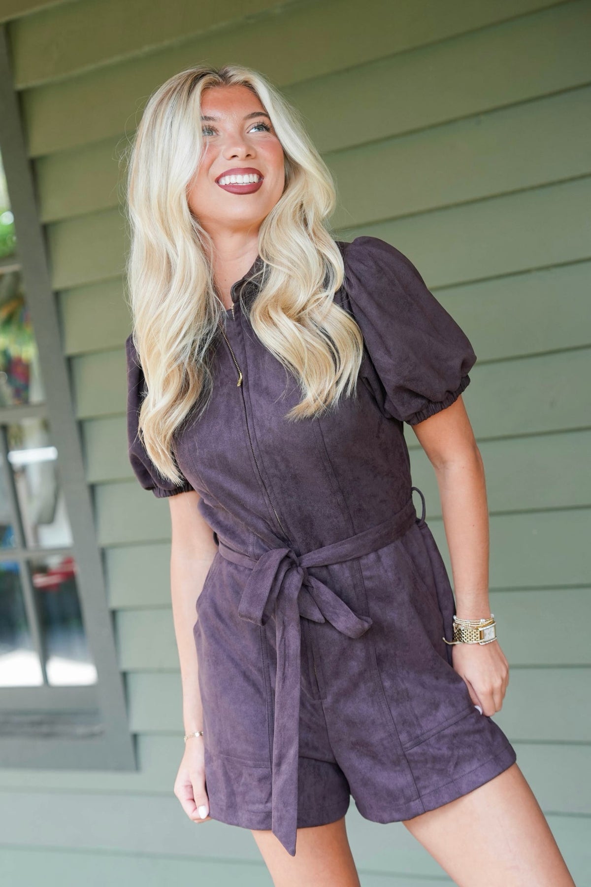 A woman with long blonde hair wearing a dark purple short-sleeve romper with puff sleeves and a tie waist, standing in front of a green house siding background.