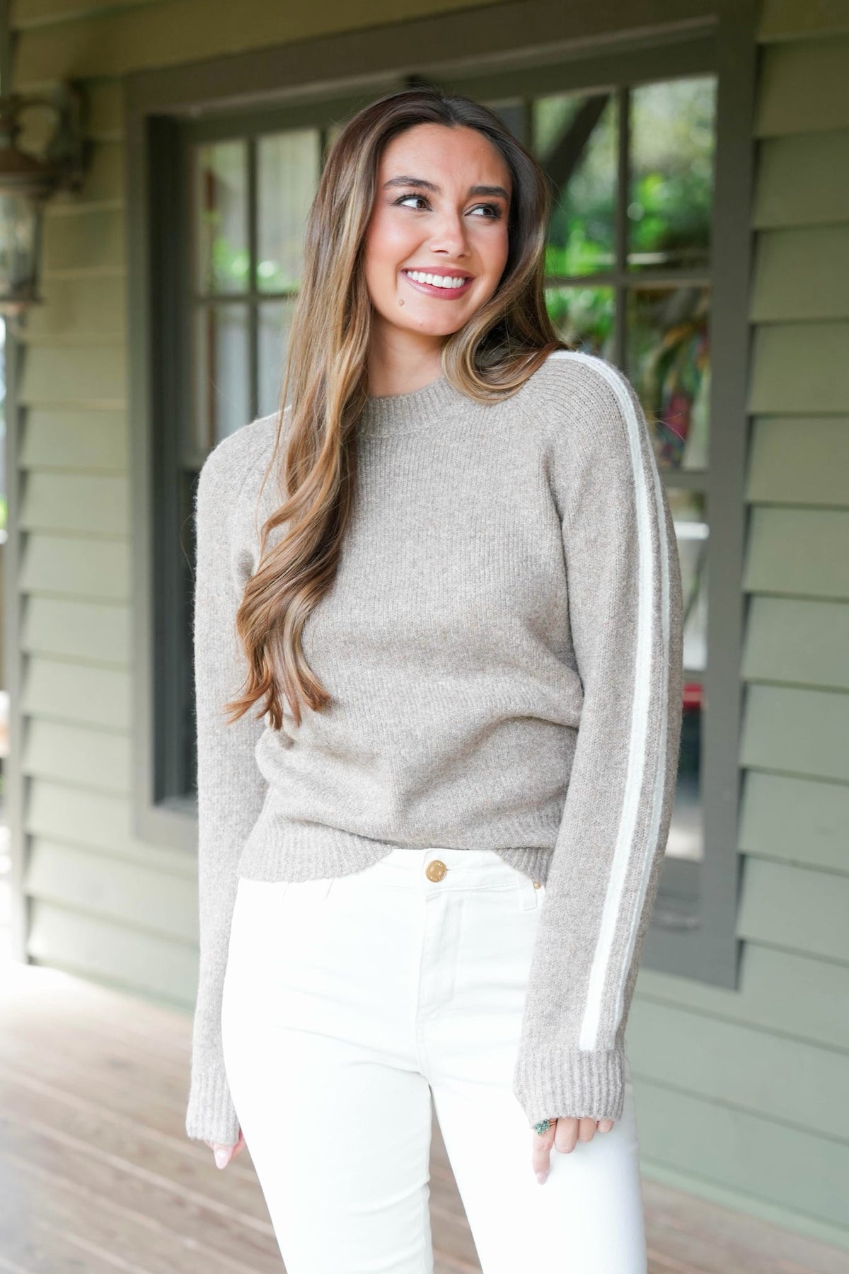 A woman wearing a light taupe knit sweater with white side stripes and white pants, standing on a porch in front of a house with green siding.