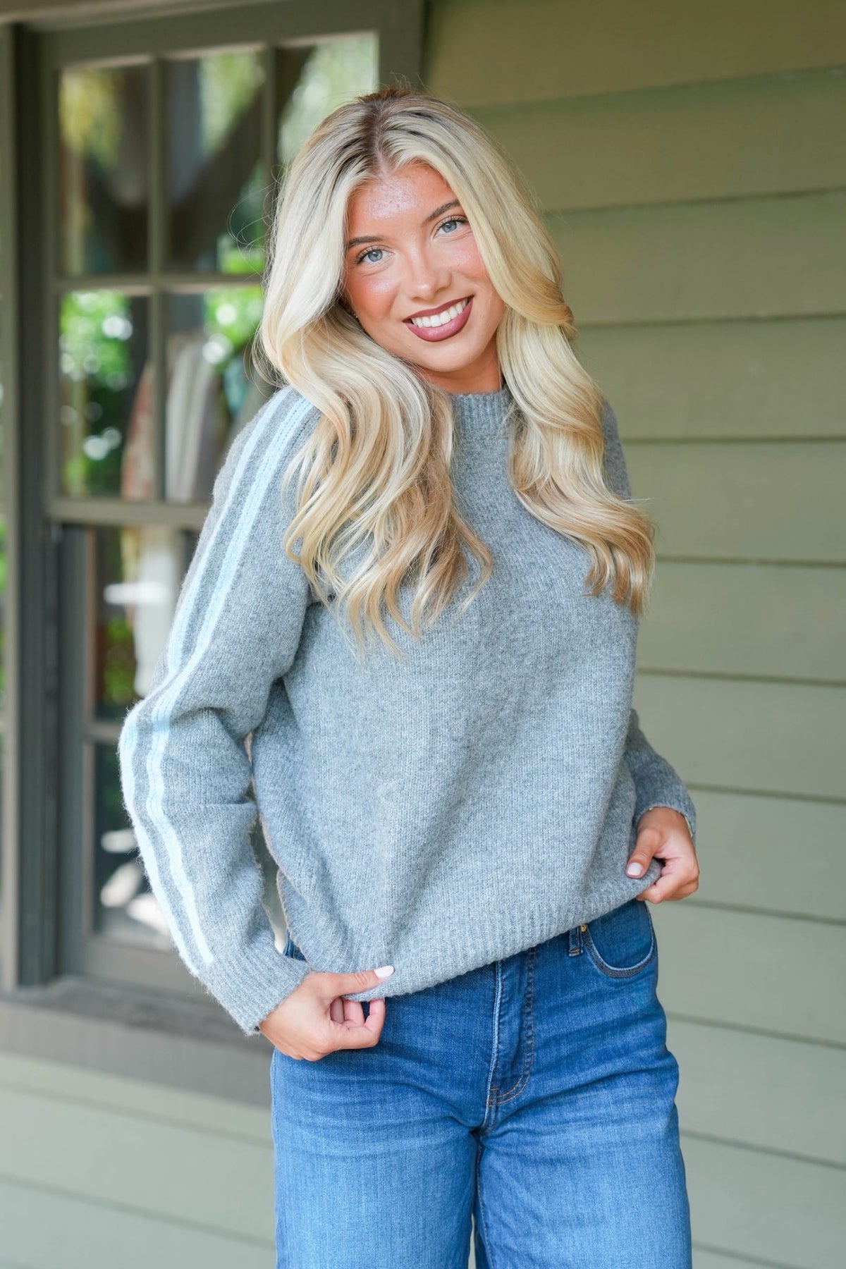 A smiling blonde woman wearing a light gray knit sweater with white stripes down the sleeves and blue jeans, standing in front of a house exterior with light green siding.