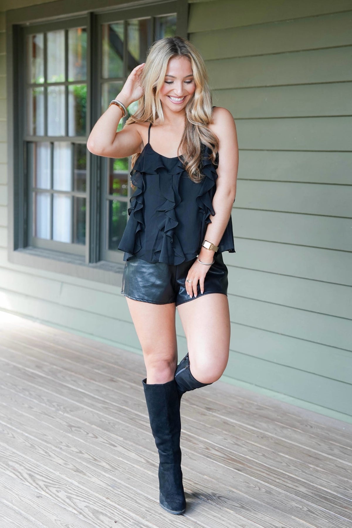 A woman posing on a wooden porch wearing a black ruffled camisole top, black faux-leather shorts, and black over-the-knee boots.