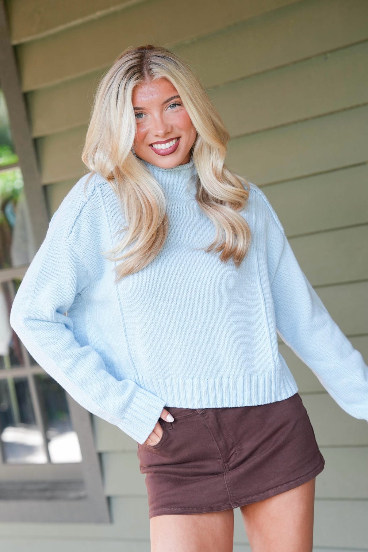 A smiling blonde woman wearing a blue ribbed knit mock-neck sweater and brown skirt, standing in front of a house exterior.