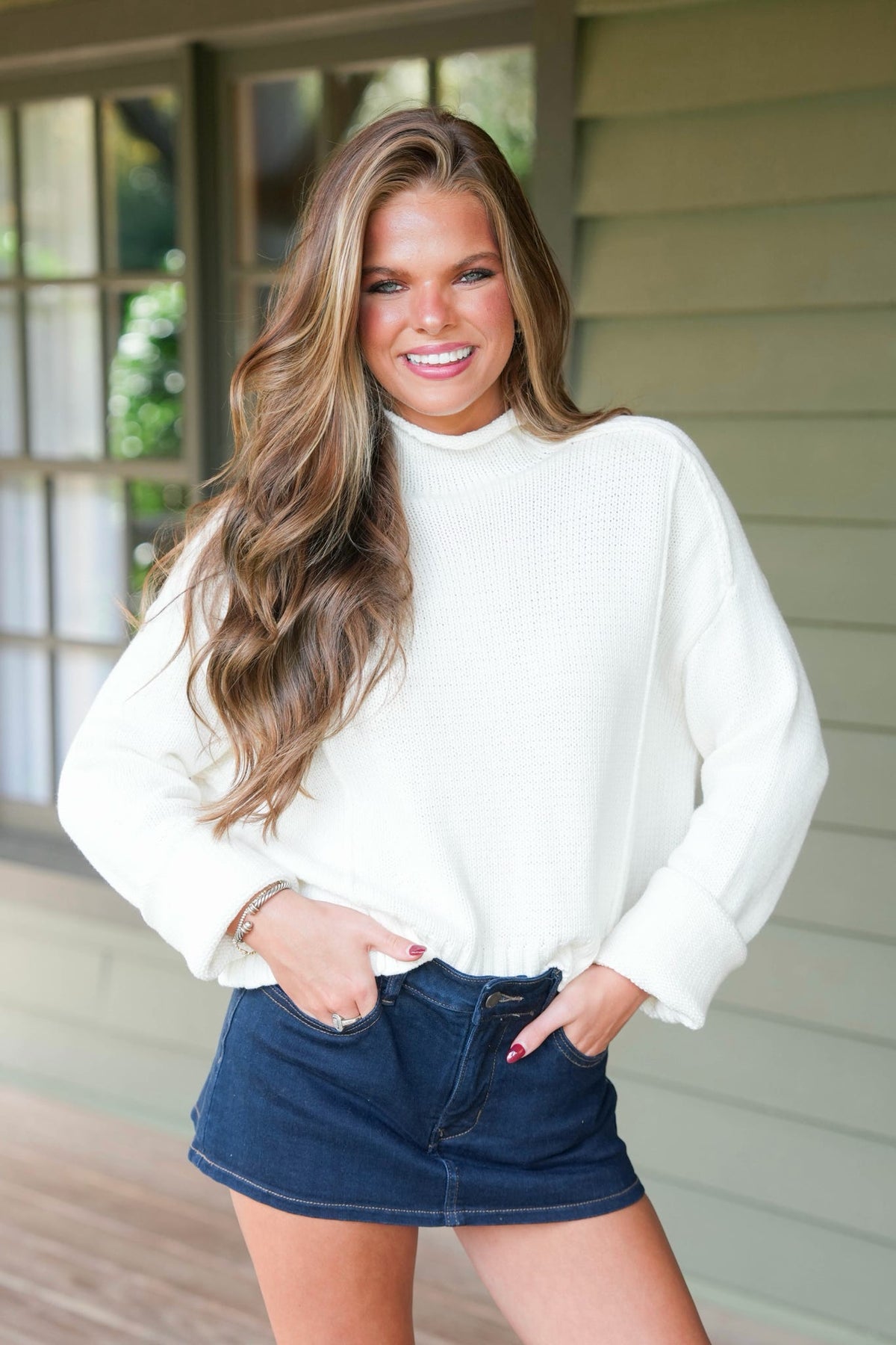 A smiling woman with long wavy brown hair stands on a porch wearing a white ribbed mock-neck long-sleeve sweater and dark blue denim shorts.