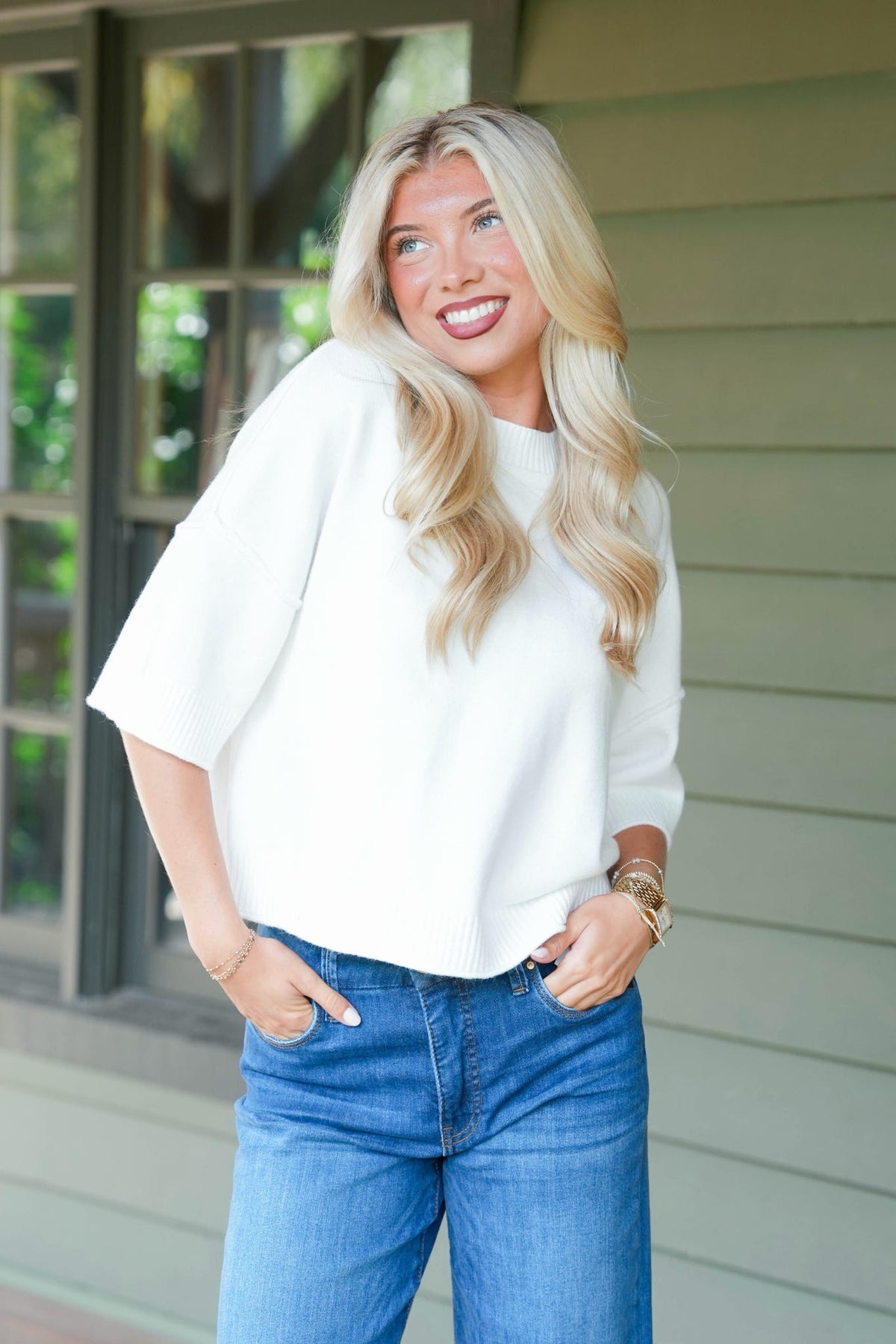 A blonde woman wearing a white short-sleeve knit top and blue jeans, standing outdoors in front of a house with light green siding and a window.