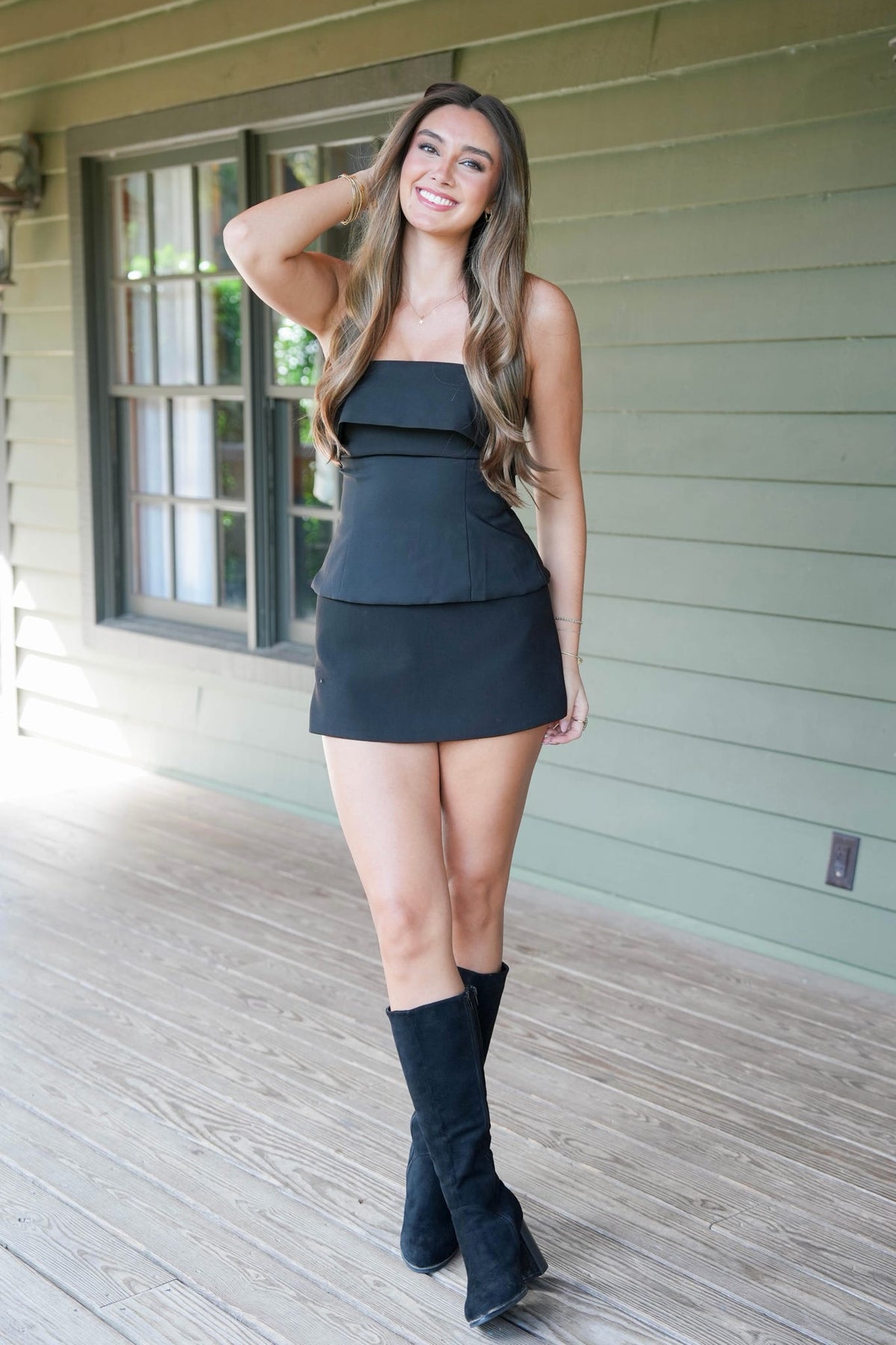 A woman with long brown hair wearing a black strapless top with a mini skirt, standing in front of a house exterior with green siding.