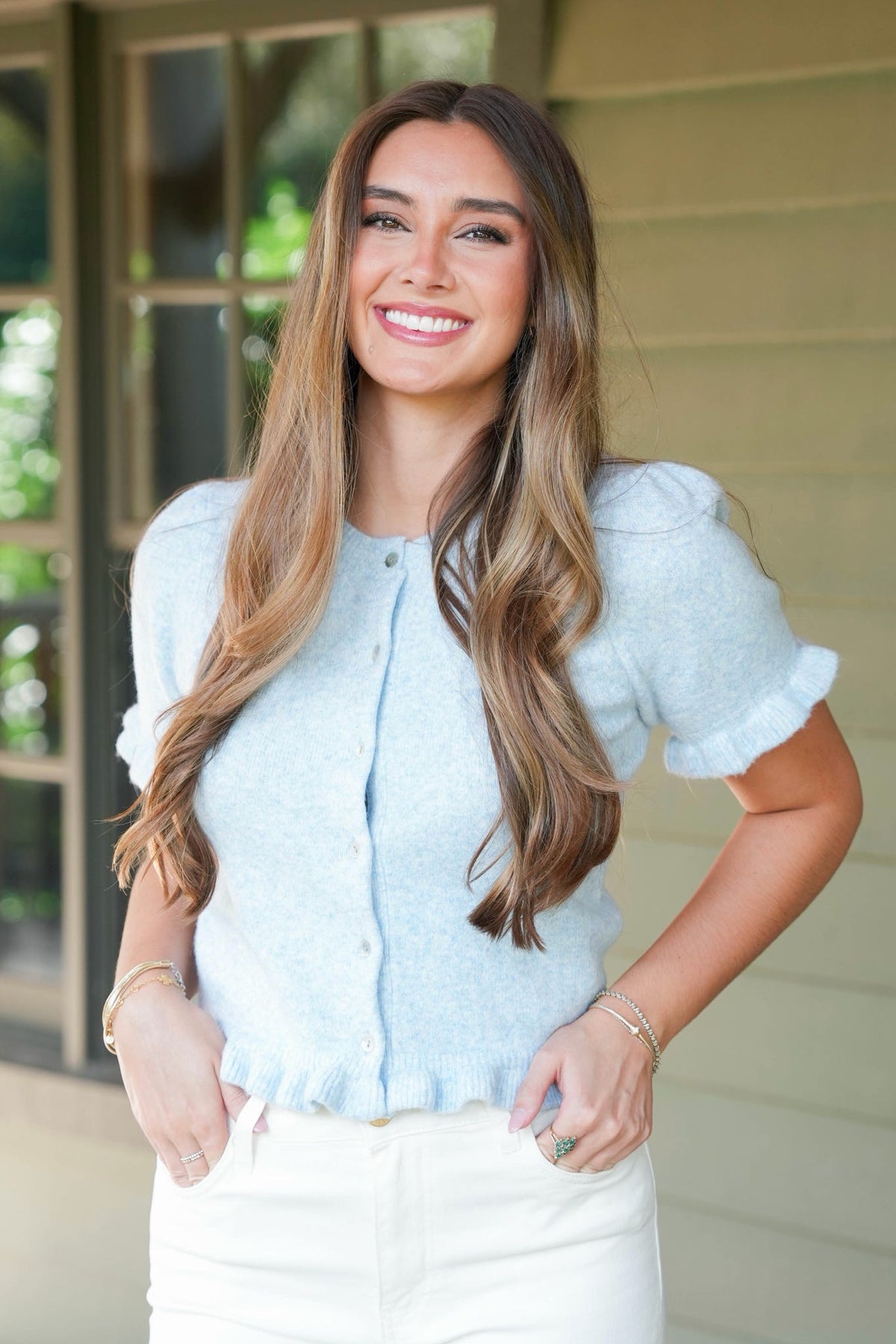 A smiling woman with long brown hair stands outdoors wearing a light blue short-sleeve knit top with ruffled trim and white pants.