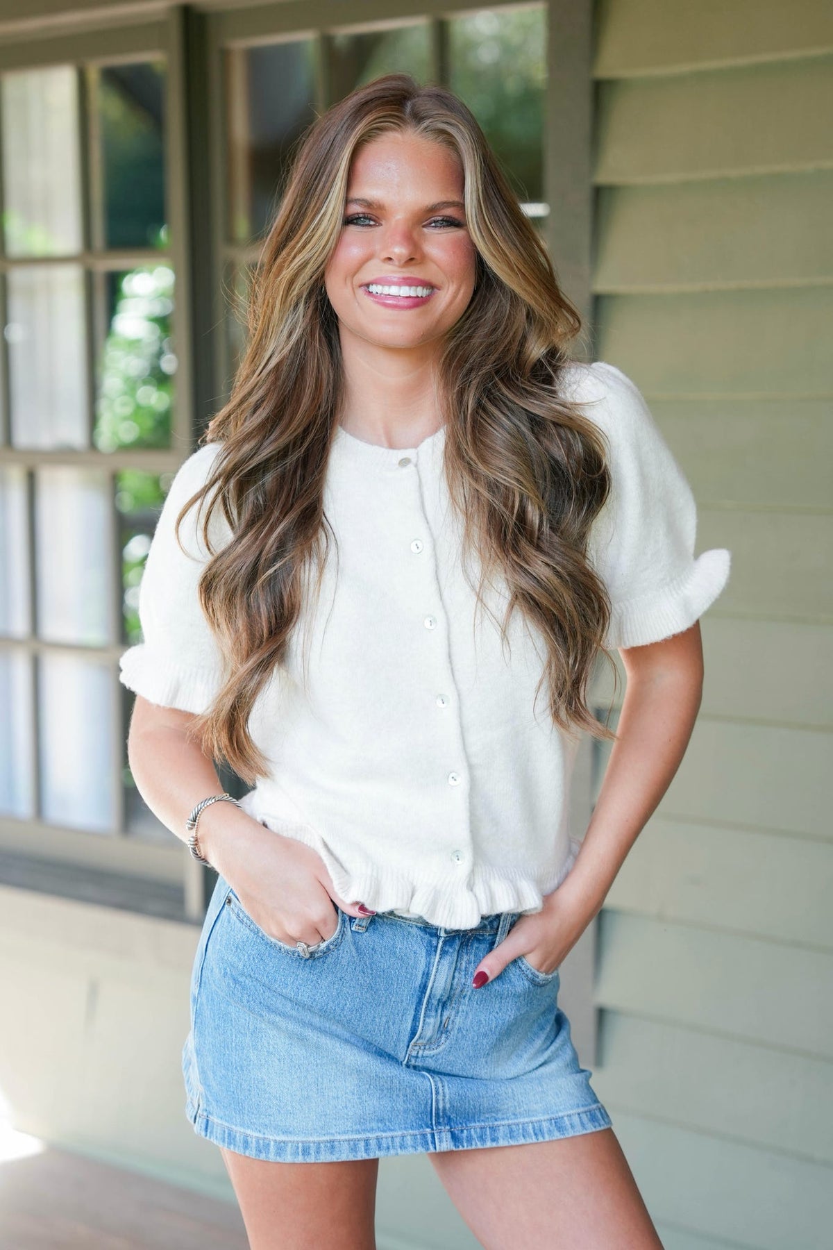 A smiling woman with long wavy brown hair stands on a porch wearing a white short-sleeve button-front blouse with ruffled cuffs and a light blue denim mini skirt.