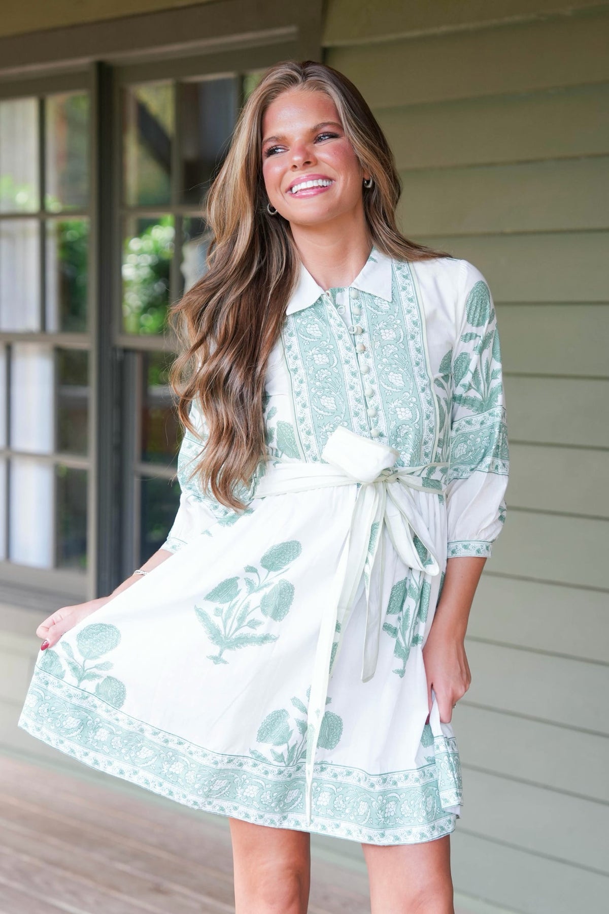 Woman standing in front of green house wearing a green and white floral dress with belted waist