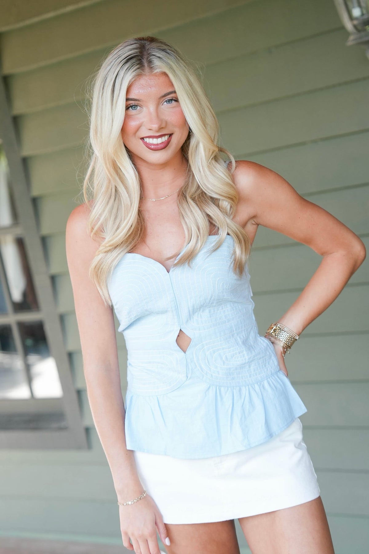 A smiling blonde woman posing outdoors in front of a house wall, wearing a light blue sleeveless peplum top with a sweetheart neckline and a white mini skirt.