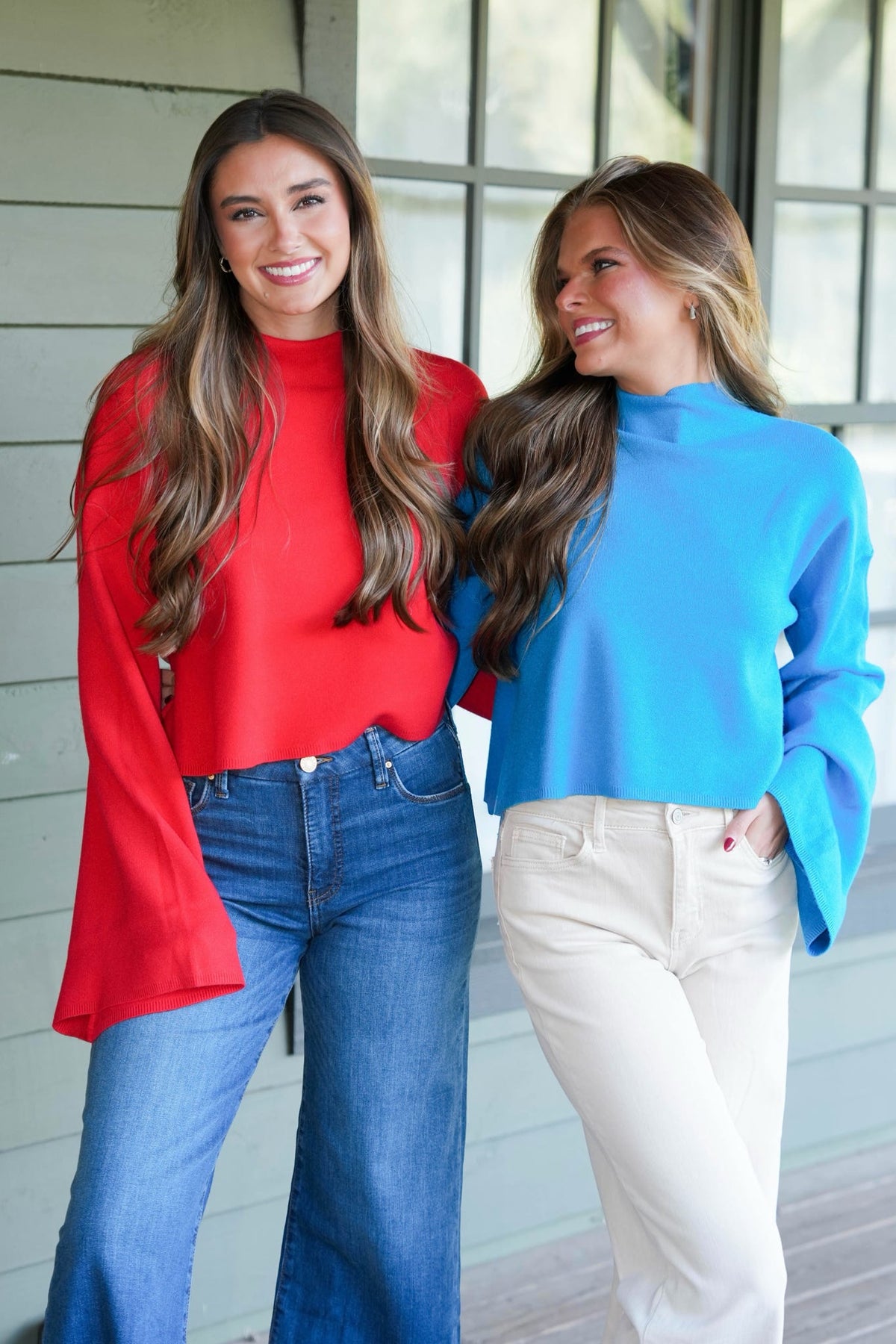 Two women wearing a bright blue mock-neck long-sleeve crop top and cream pants and a red mock-neck long-sleeve and denim pants, standing in front of a modern house exterior with large windows.