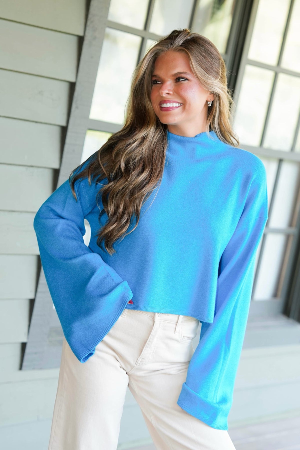 A woman wearing a bright blue mock-neck long-sleeve crop top and cream pants, standing in front of a modern house exterior with large windows.