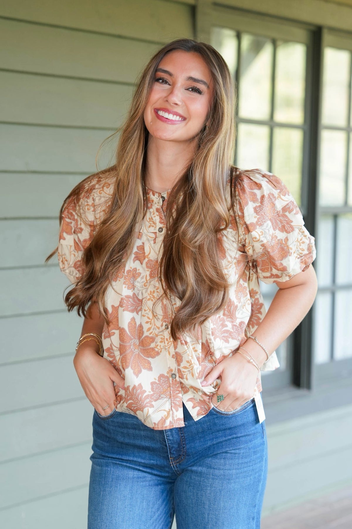 A smiling woman with long wavy brown hair stands on a porch wearing a short-sleeve button-up blouse with a beige, brown, and orange floral pattern, paired with blue jeans.