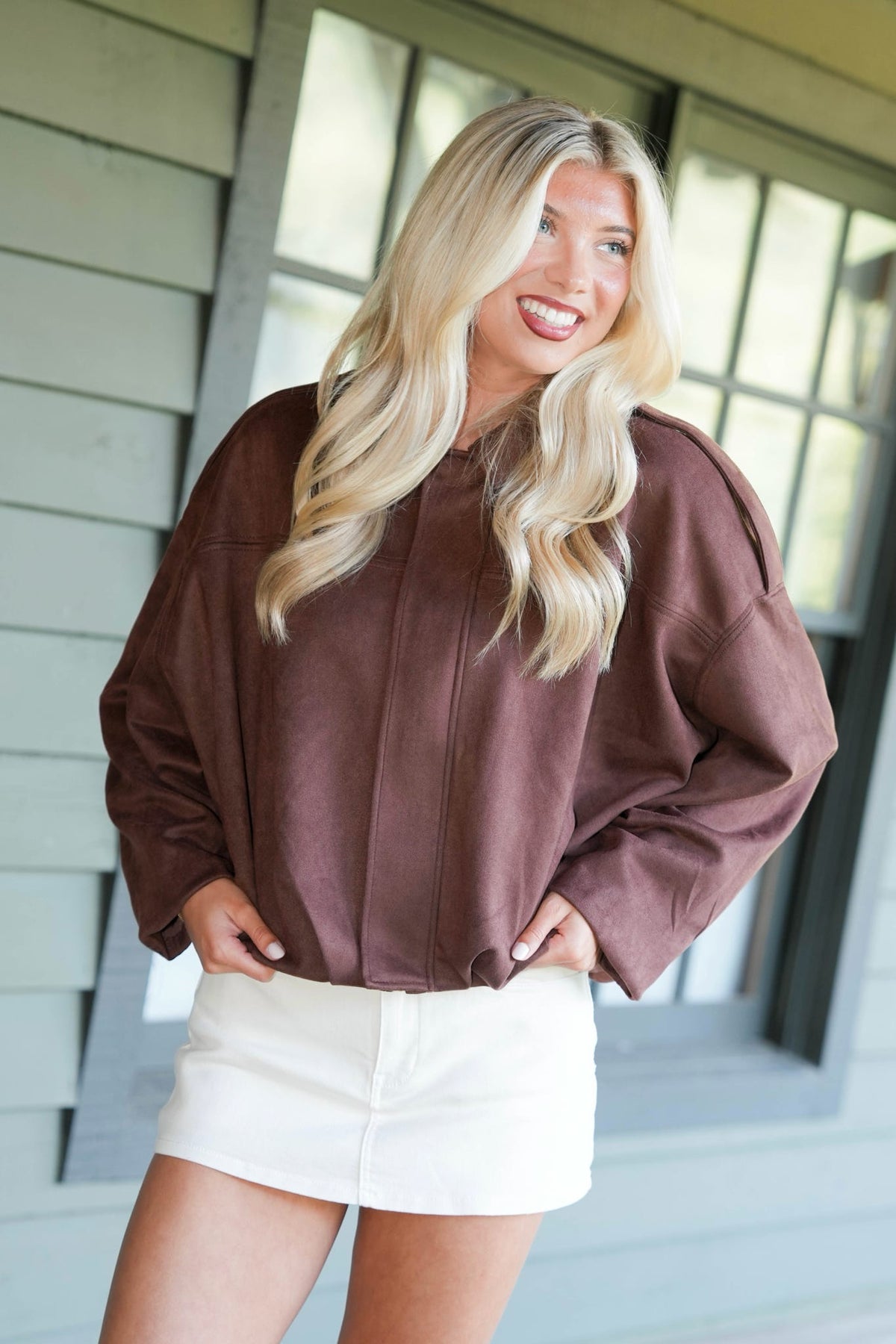 A smiling blonde woman wearing a brown oversized long-sleeve top and white shorts, standing in front of a house exterior with siding and windows.