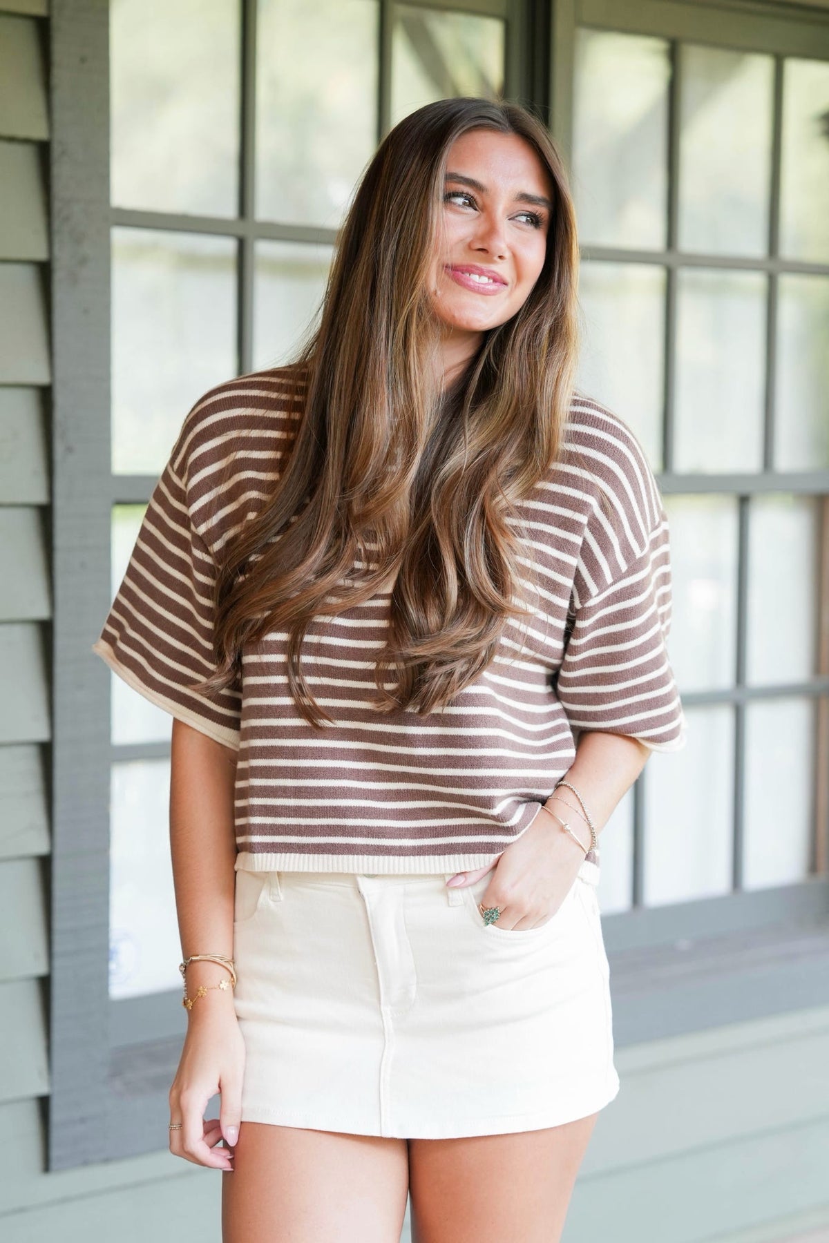 A woman with long brown hair stands in front of a window wearing a brown and white horizontally striped short-sleeve top and a white mini skirt.