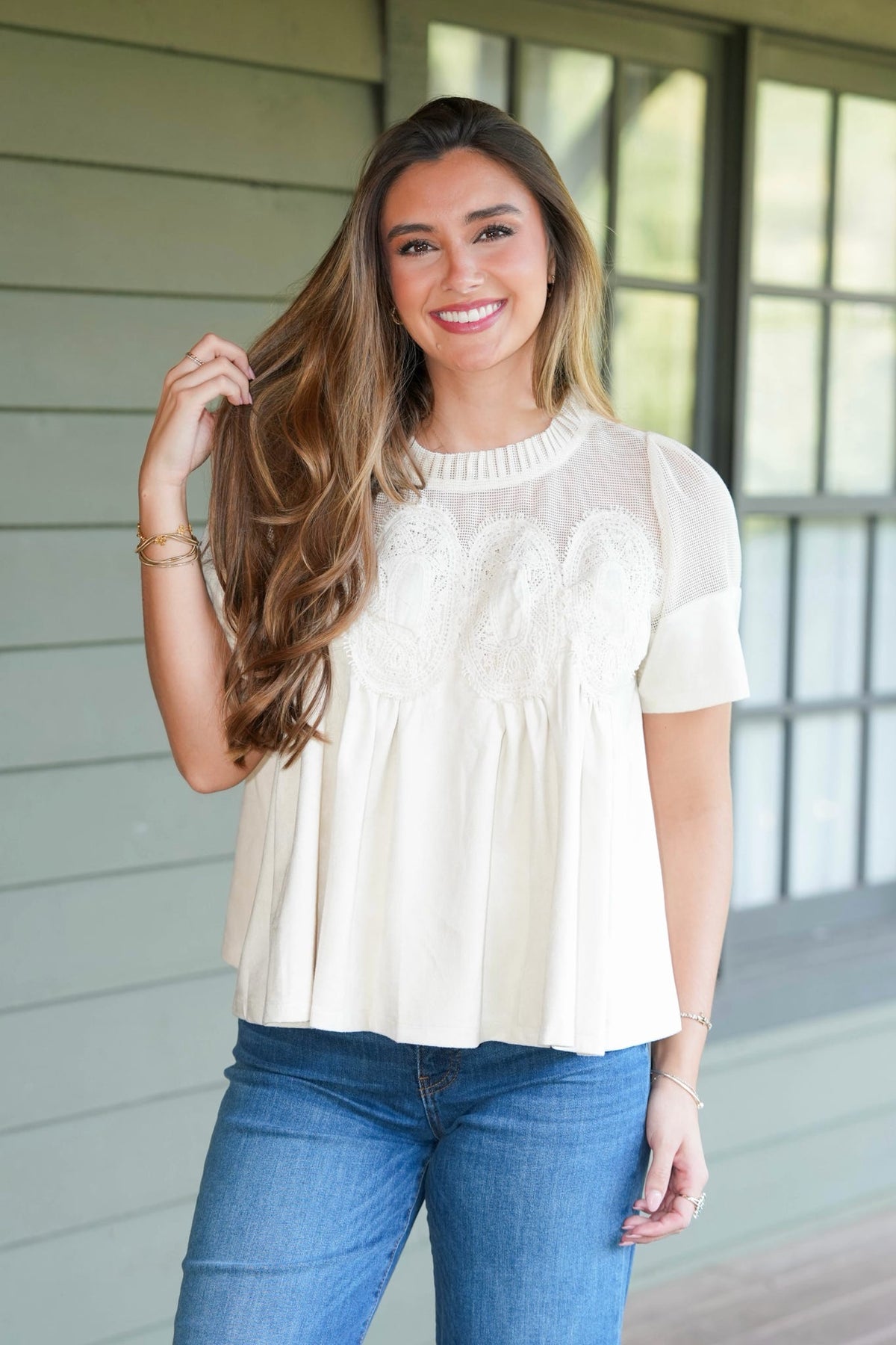 A woman wearing a cream-colored short-sleeve blouse with a sheer mesh yoke and embroidered chest detail, paired with blue jeans, standing in front of a house porch.