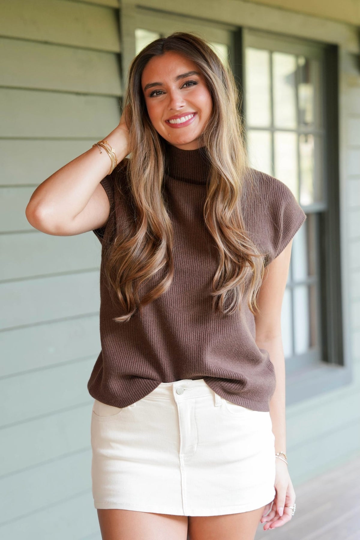 A woman with long wavy brown hair stands on a porch wearing a brown ribbed mock-neck top and an off-white mini skirt.
