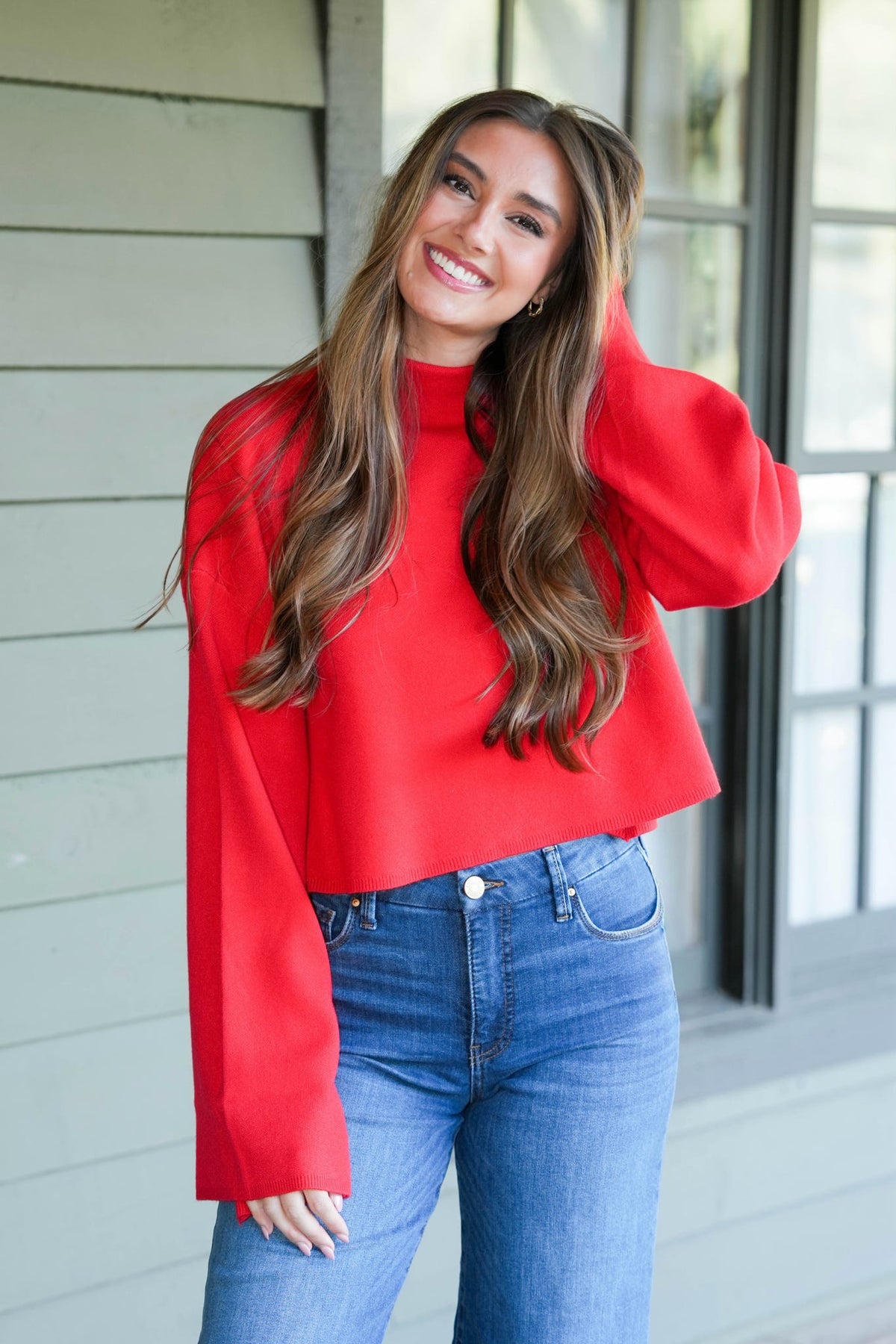 A woman wearing a bright red mock-neck long-sleeve crop top and blue jeans, standing in front of a light gray siding wall with a window.