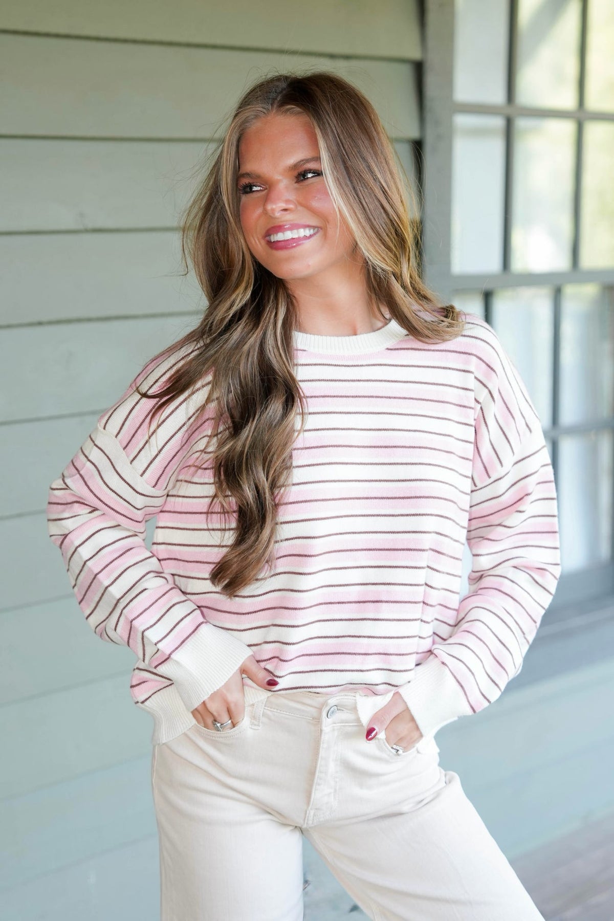 A woman with long brown hair smiling and looking to the side, wearing a pink and white striped long-sleeve crewneck sweatshirt and cream-colored pants, standing in front of a light gray wood-paneled wall.