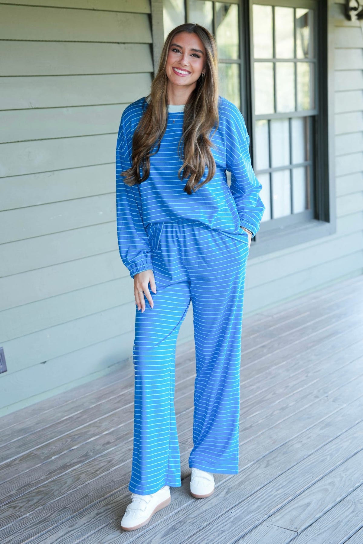 A woman modeling a matching blue and white striped lounge set consisting of a long-sleeve top and wide-leg pants, standing on a wooden porch.