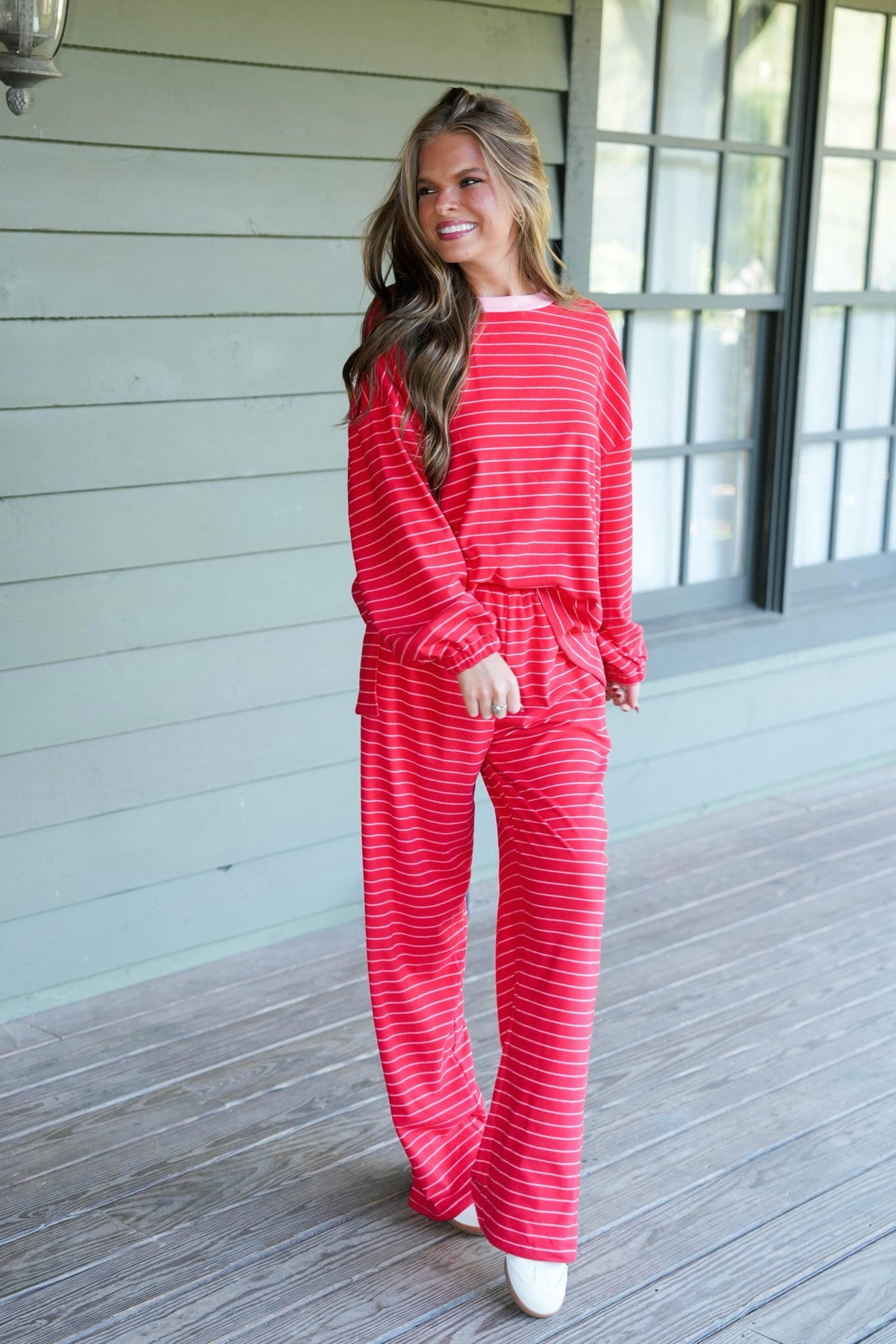 A woman modeling a matching red and white striped lounge set consisting of a long-sleeve top and wide-leg pants, standing on a wooden porch.