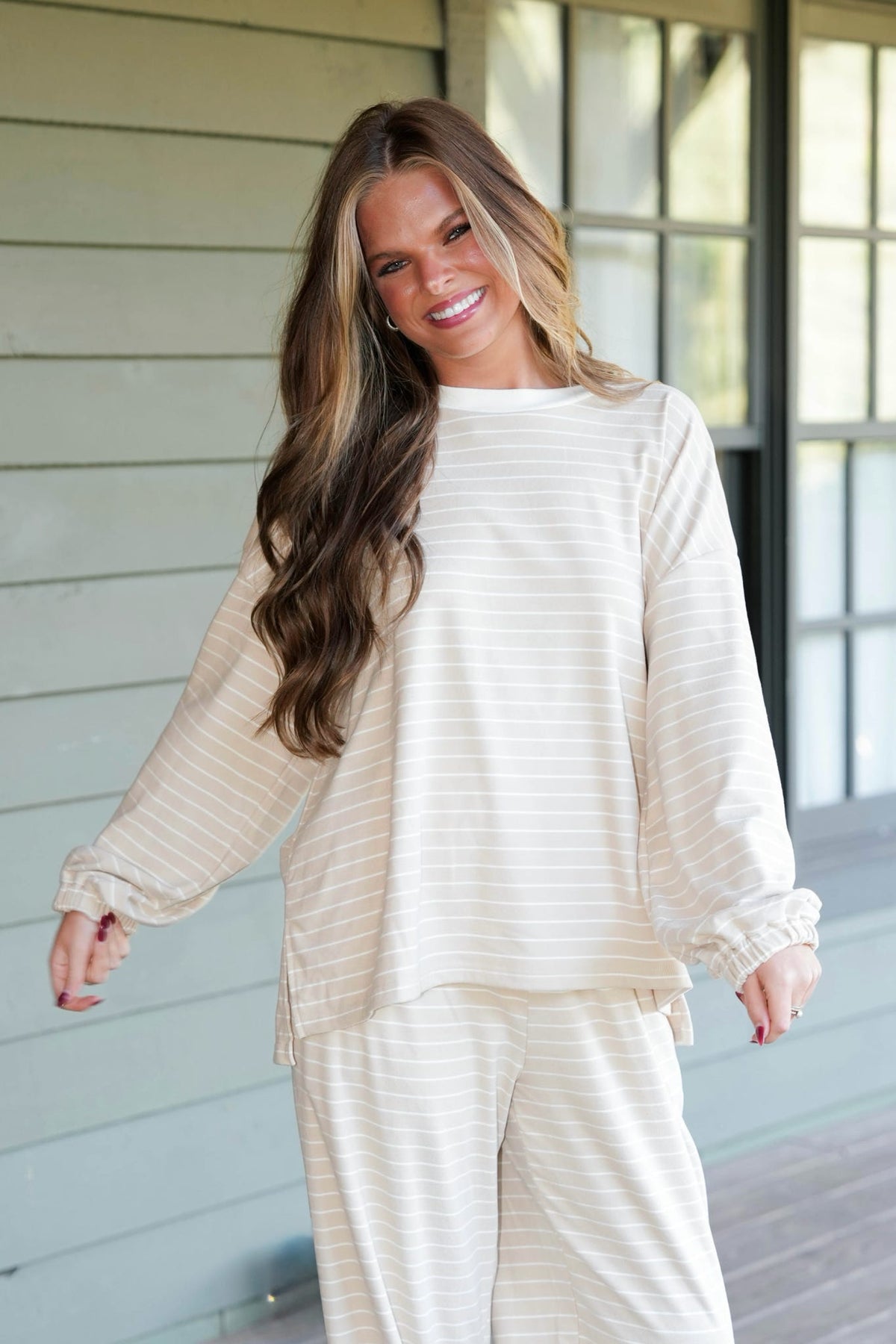 A woman modeling a matching two-piece loungewear set consisting of a long-sleeve top and pants, both in a cream and white stripe pattern, standing on a porch.