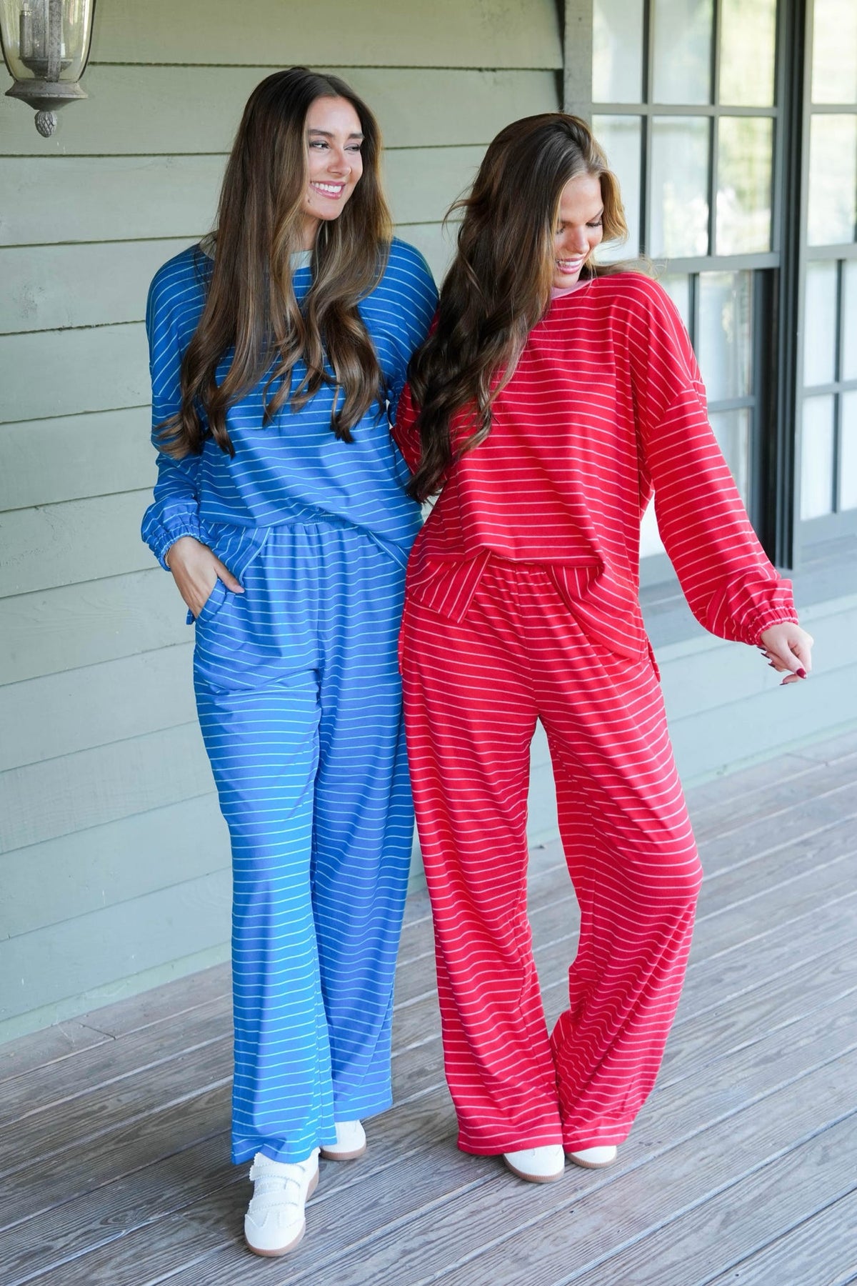 Two women modeling matching lounge sets on a porch. One wears a blue and white striped long-sleeve top and matching pants, and the other wears a red and white striped long-sleeve top and matching pants.