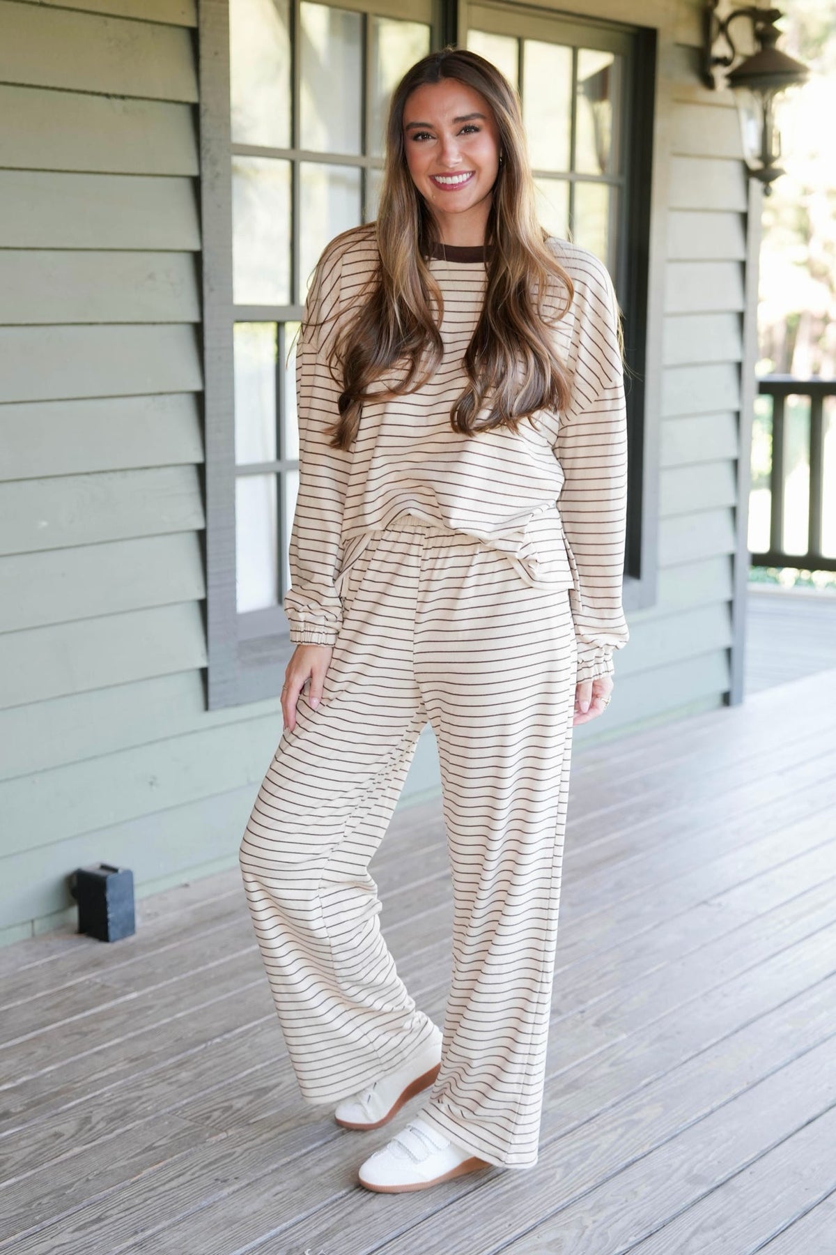A woman stands on a wooden porch wearing a beige and white striped long-sleeve jumpsuit with a drawstring waist and matching wide-leg pants, paired with white sneakers.