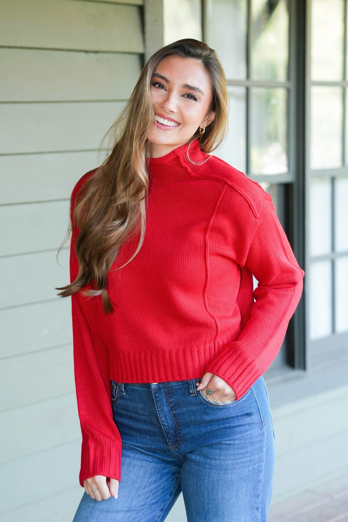 A woman wearing a bright red ribbed mock neck sweater and blue jeans, standing in front of a light-colored house exterior.